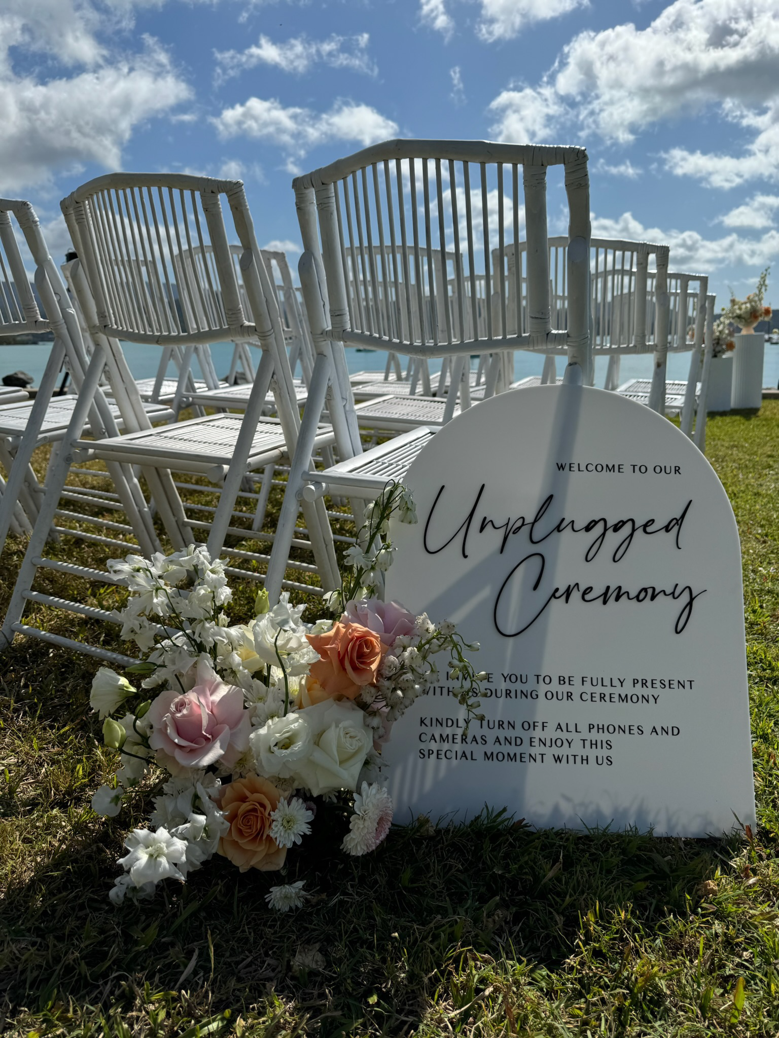 Outdoor wedding setup with white chairs on grass, floral arrangements, and a sign that says 'Welcome to our Unplugged Ceremony' against a lakeside backdrop with blue sky and clouds.