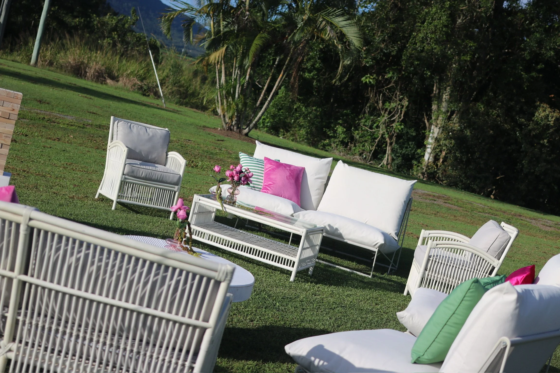 Outdoor patio furniture set on a grassy lawn with pink, green, and striped cushions, a small floral centerpiece, and surrounded by lush green trees.
