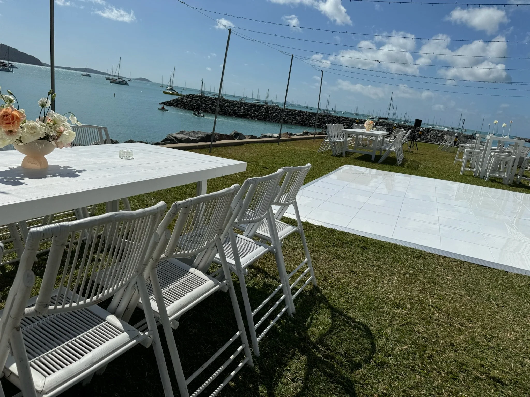 Outdoor event setup with white tables and chairs, floral centerpiece, overlooking a marina with sailboats, and a clear sky