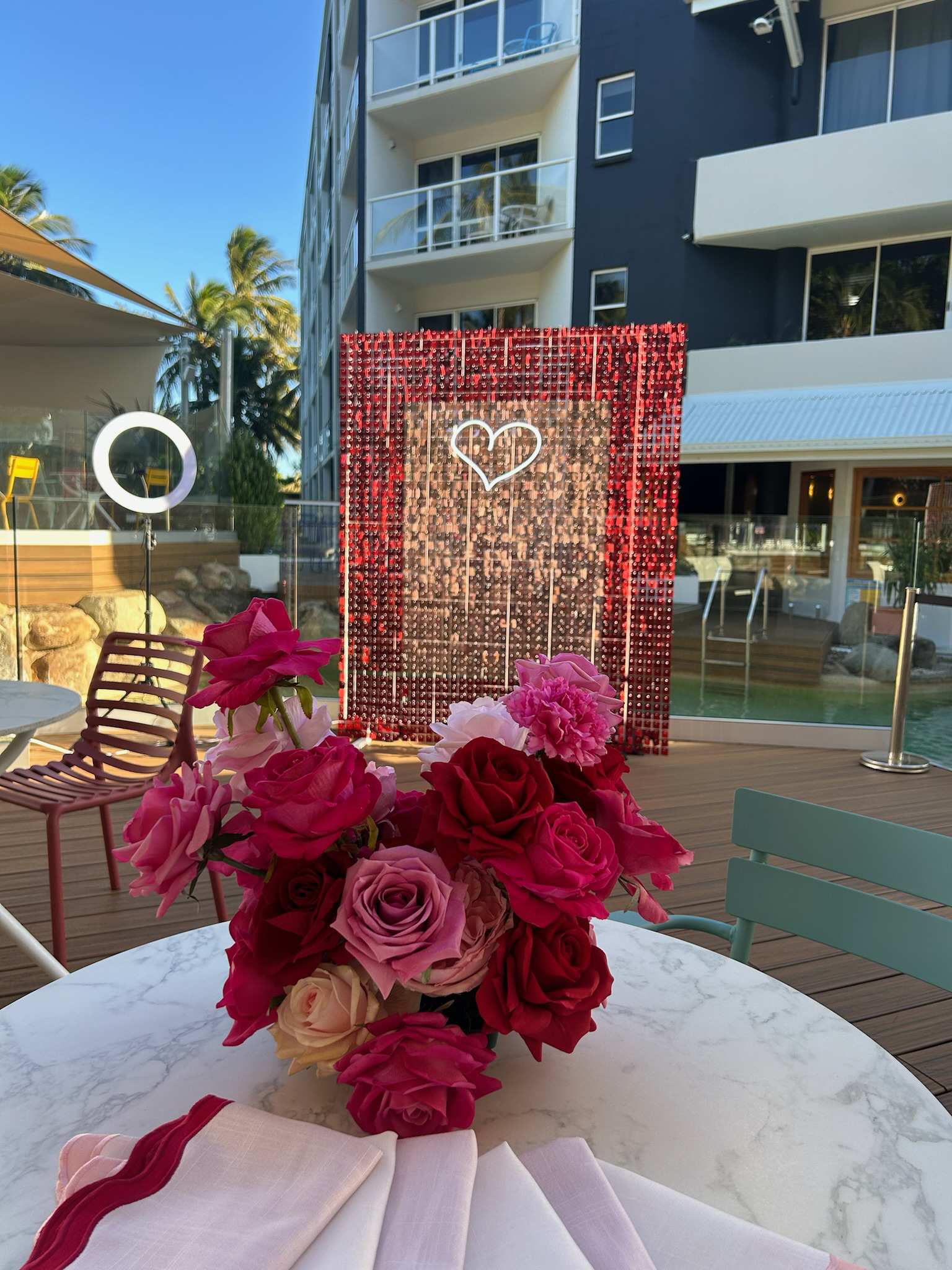 A floral arrangement of pink, red, and cream roses on a white marble table. In the background, a decorative backdrop with a glowing heart outline and hanging red beads, set outdoors near a pool area with modern building and palm trees.