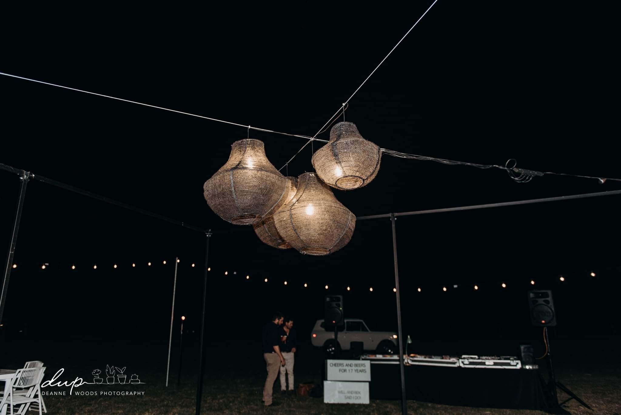 A nighttime outdoor event with four hanging woven or rattan lanterns illuminated overhead, and a small gathering of people in the background near a sign that reads 'Cheers and Beers for 17 Years' and musical equipment.