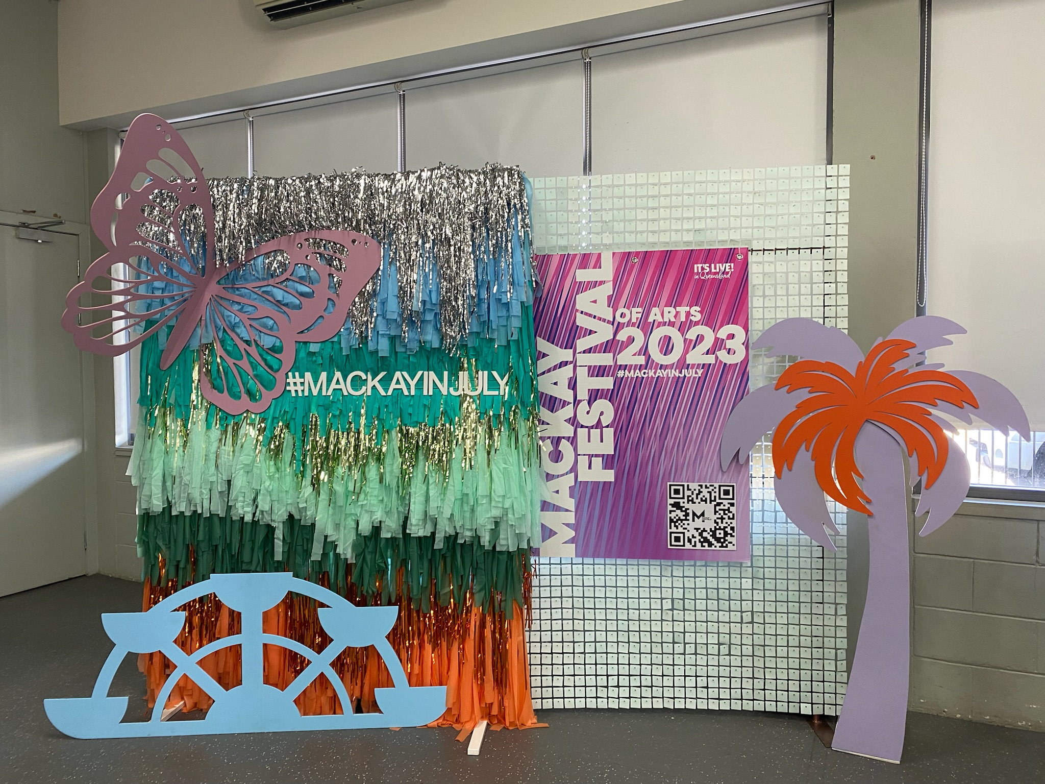 Decorative display for Mackay July Arts Festival 2023, including a colorful tassel backdrop, a pink butterfly cutout, a purple and orange palm tree, a blue boat cutout, a poster with event details, and metallic streamers.