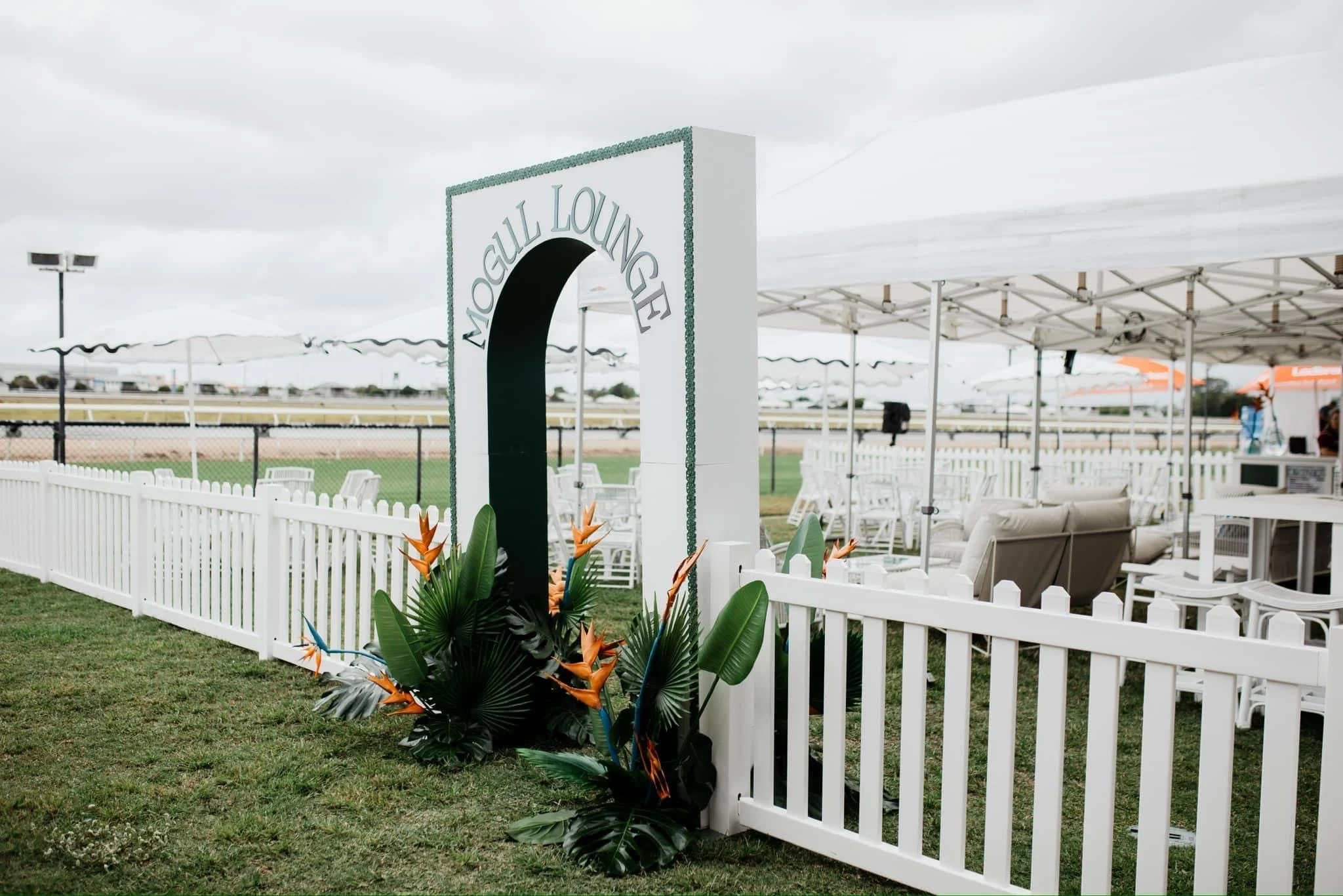 Decorative sign reading 'Mogull Lounge' with tropical foliage, set up in an outdoor area with white fencing and seating under canopy tents.