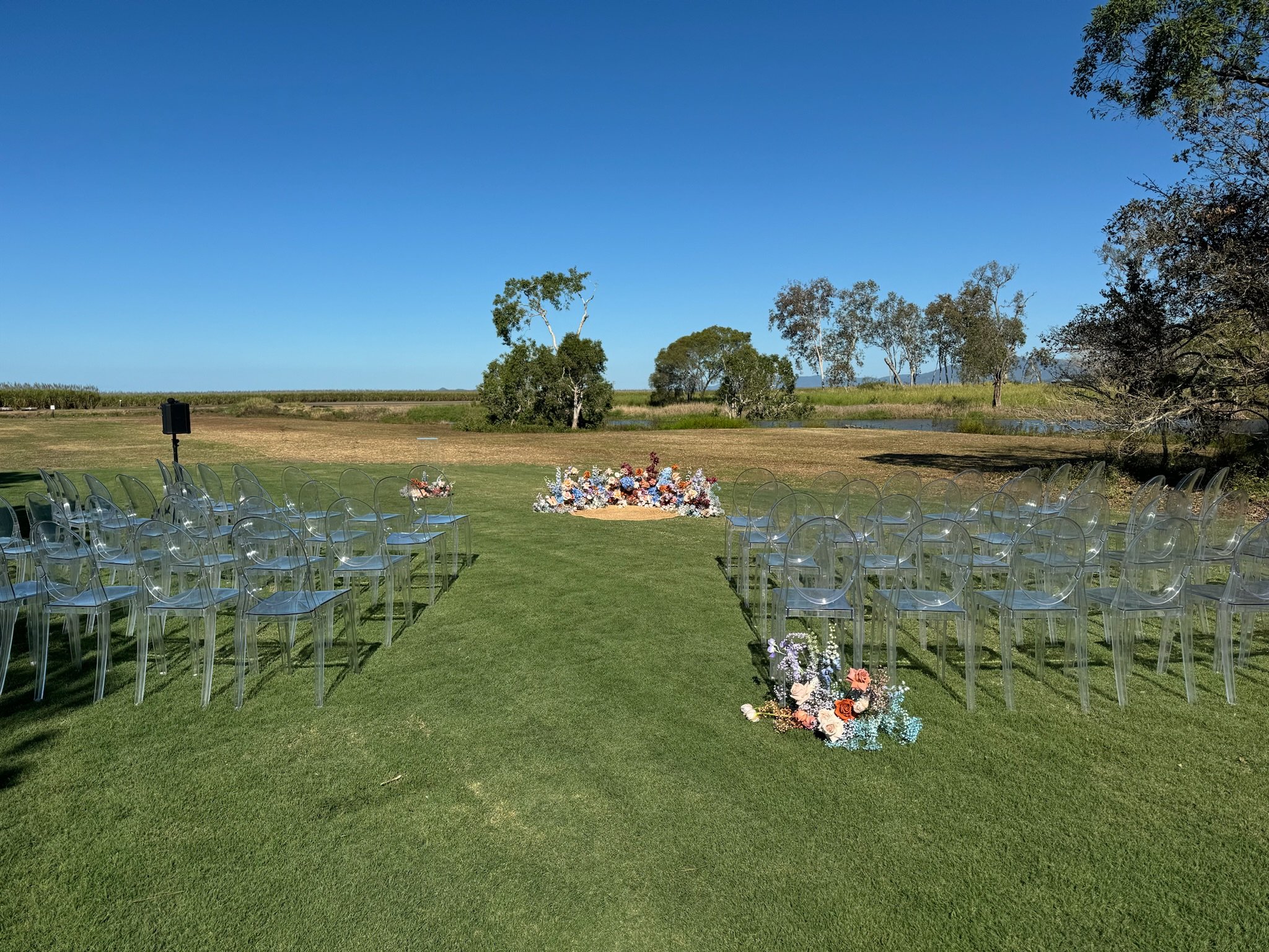 Outdoor wedding setup with rows of transparent chairs on a grassy field, decorated with floral arrangements along the aisle, with a scenic view of trees, water, and blue sky in the background.