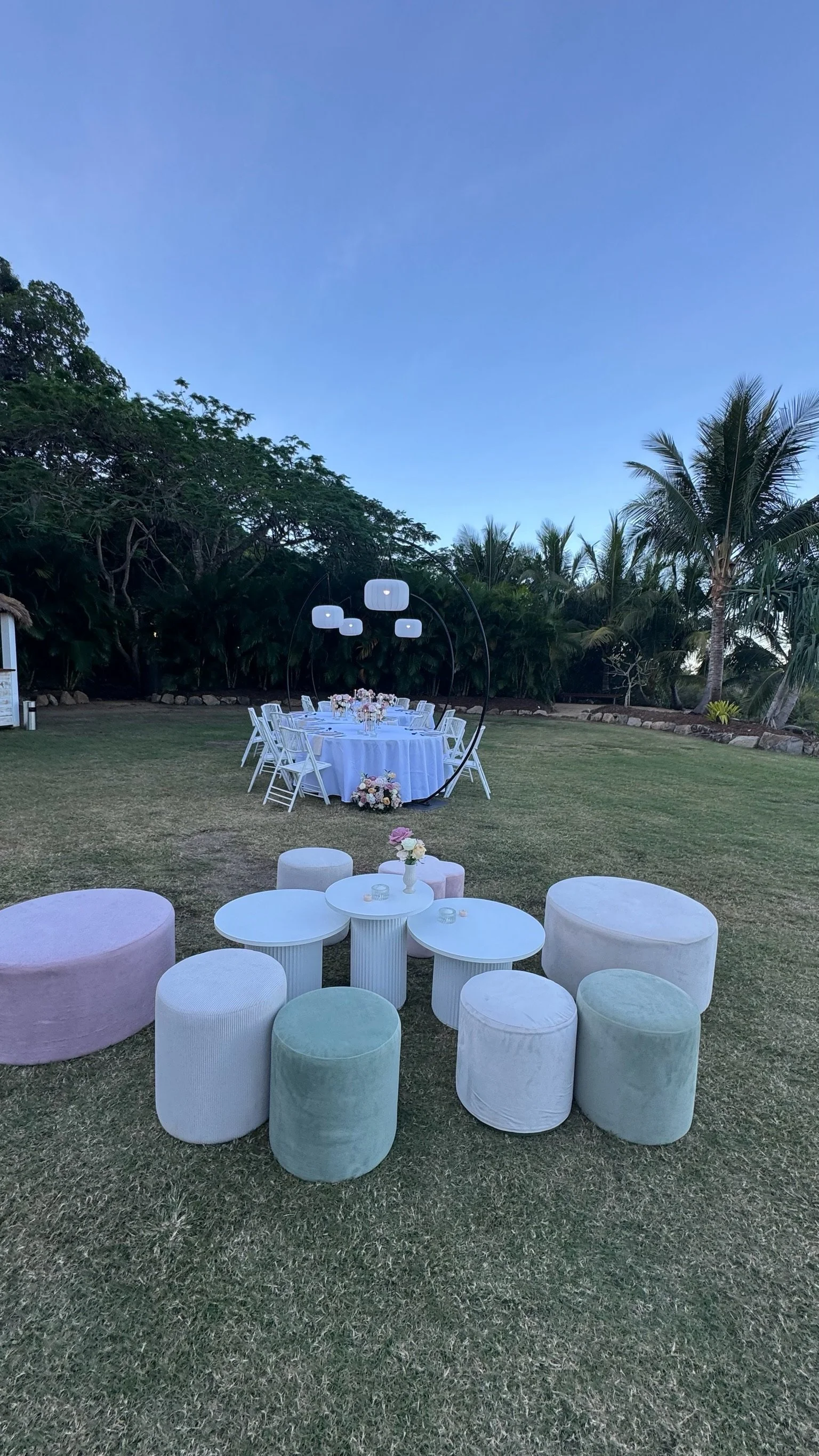 Outdoor wedding setup with a round table with white chairs, floral arrangements, and modern hanging lights, all on a grassy area with trees and palm trees in the background.