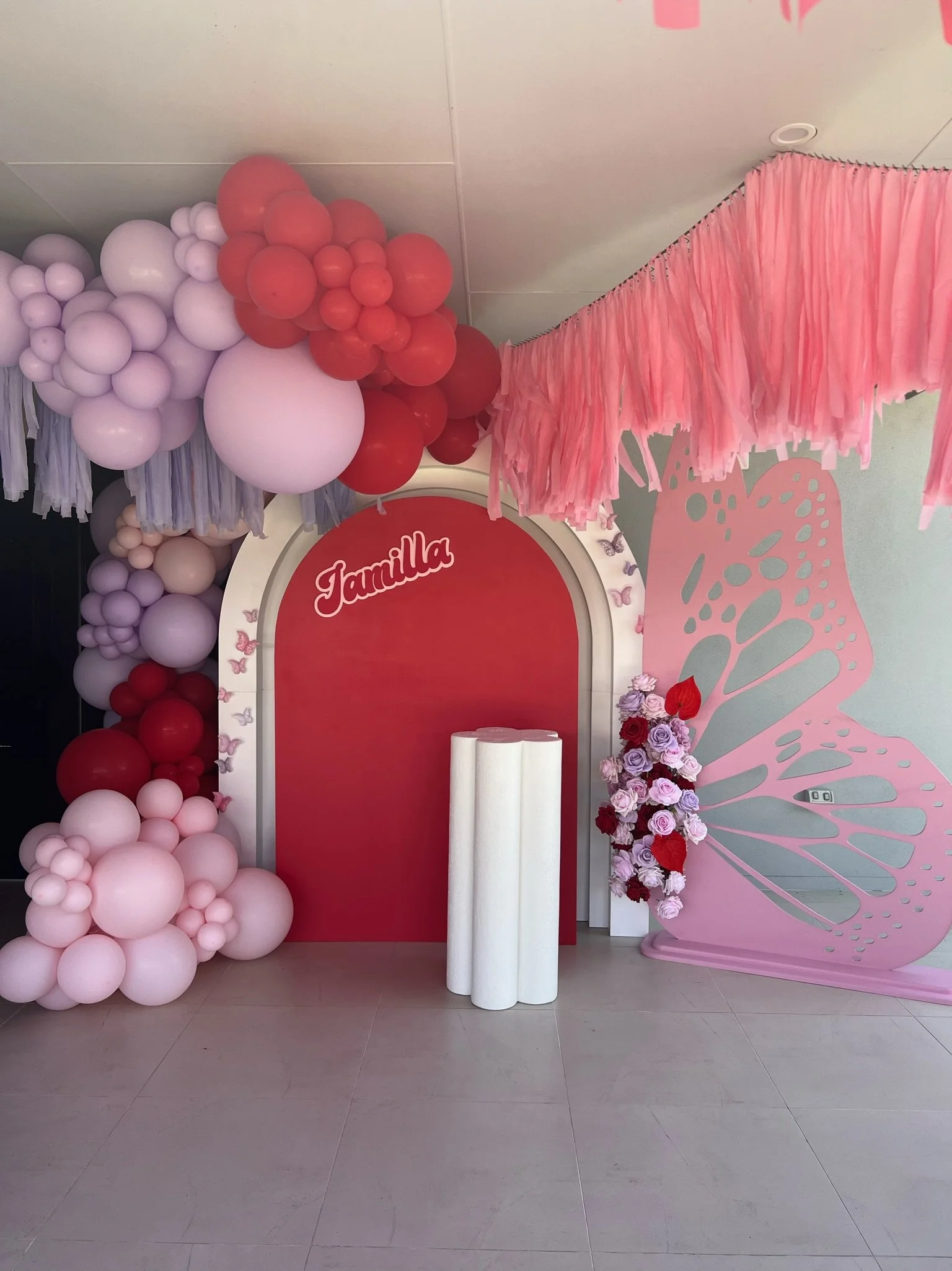 Decorative birthday party backdrop with pink and red balloons, pink paper fringe hanging from ceiling, a large pink butterfly cutout on the right, and a floral arrangement with pink and red roses on the right side of the pink arch with the name 'Jami