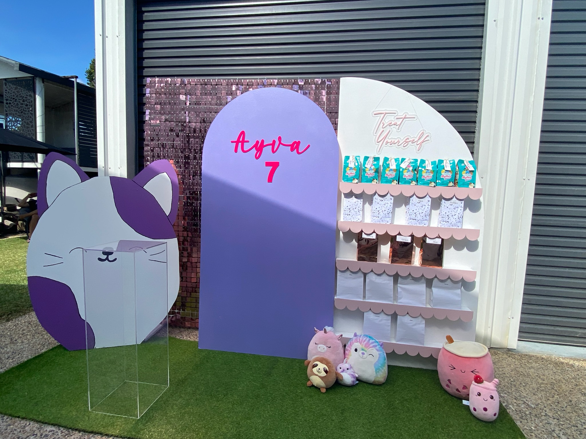 Decorative display for a 7th birthday celebration with a purple and white cat face cutout, purple and white themed backdrop with the name 'Ayyva' and the number '7', plush toys, and a small shelf with snack packages.