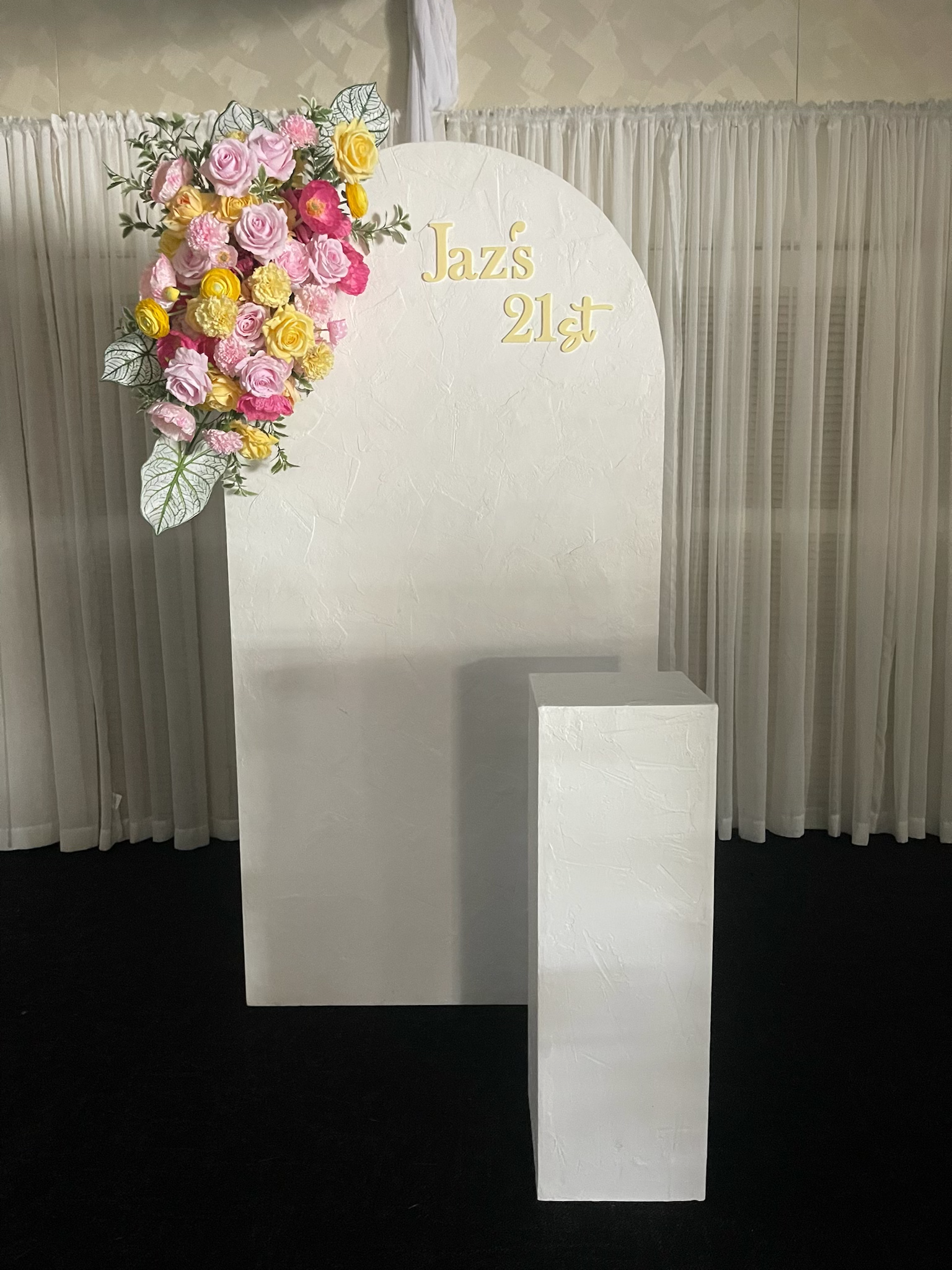 A floral decoration with pink, yellow, and white flowers on a white backdrop with the text "Jaz's 21st" in gold lettering, likely for a 21st birthday celebration.
