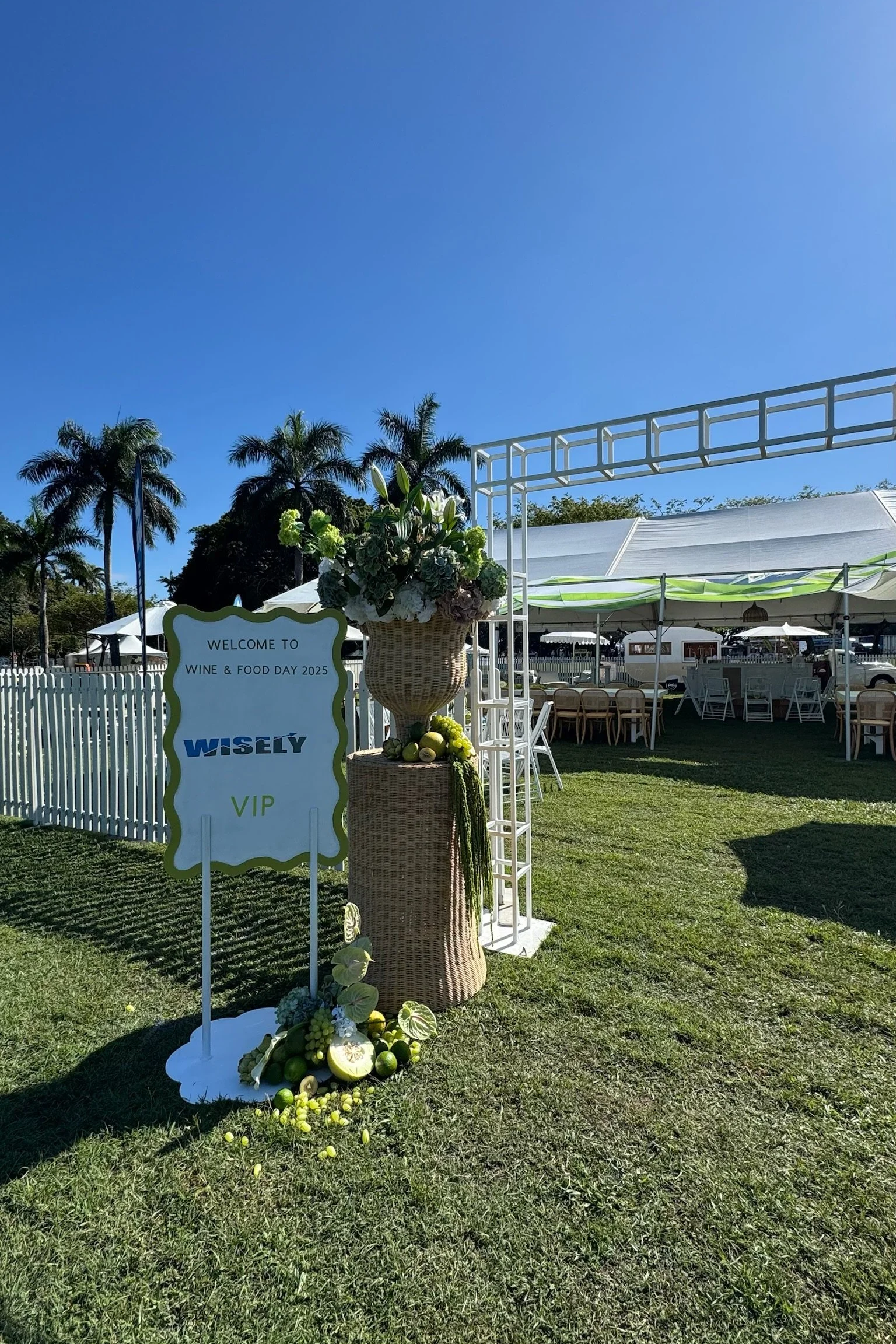 Outdoor event setup with white tent, palm trees, floral arrangements, and a sign welcoming guests to Wine & Food Day 2025, VIP section.