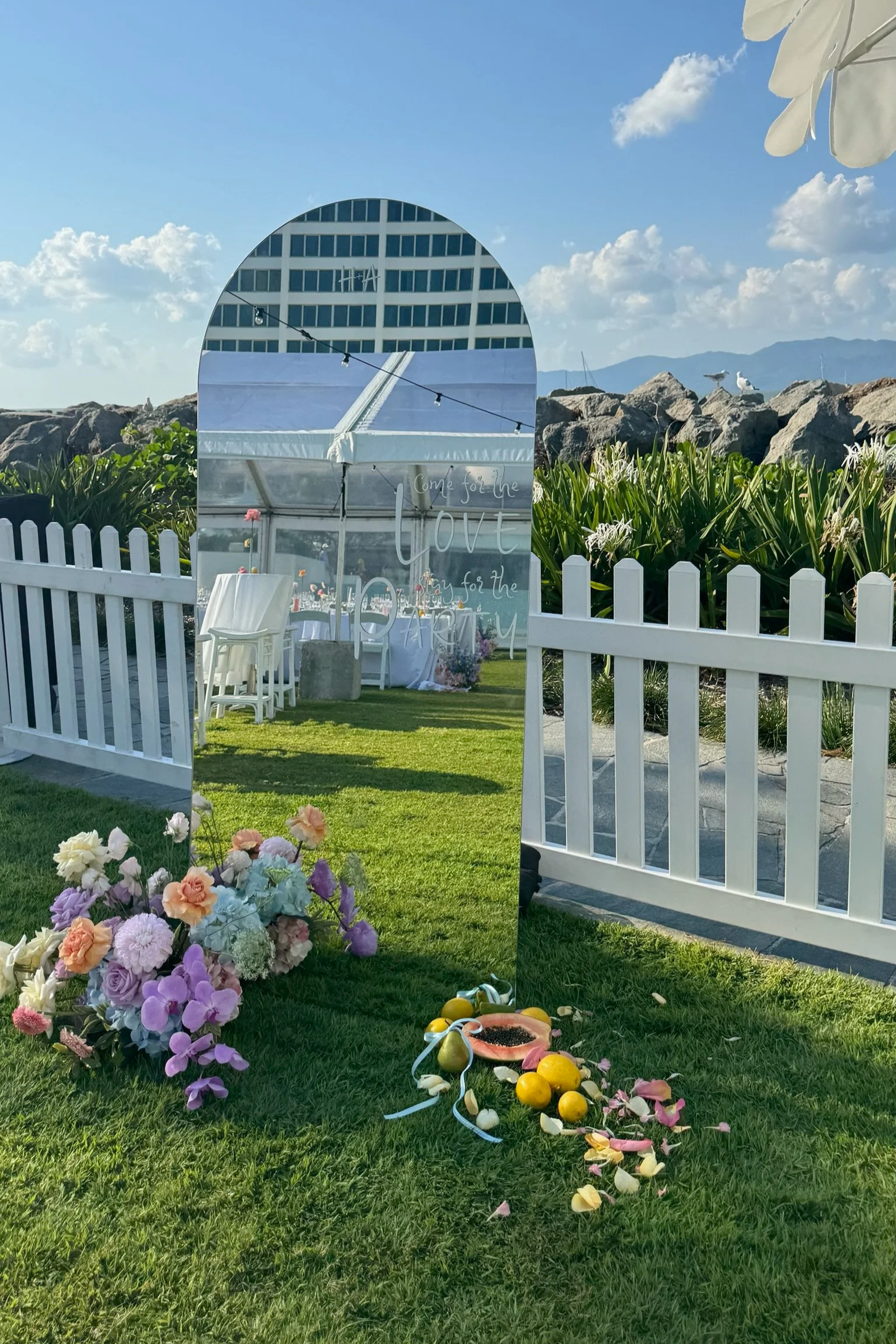A decorated outdoor party setup with a mirror displaying handwritten white text, pink and purple flowers on the grass, a white picket fence, a tent in the background, and a scenic mountain view with blue sky and clouds.