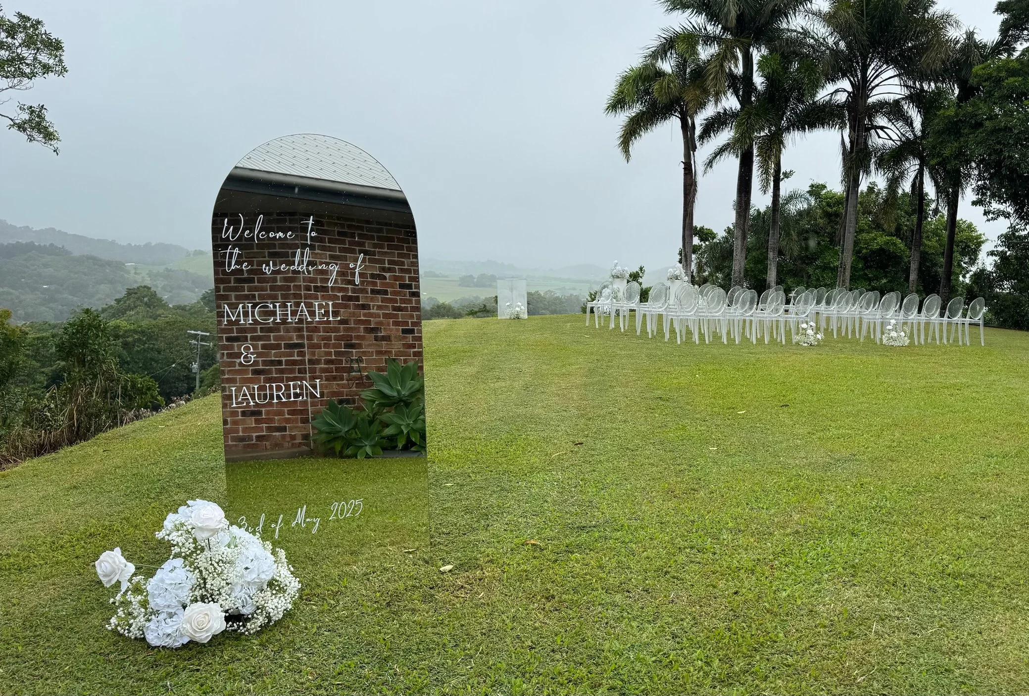 Outdoor wedding setup with transparent chairs arranged in a row on a grassy hill, with a decorative floral arrangement at the front and a tall mirror with wedding details inscribed in cursive. Behind are tall palm trees and a cloudy sky.