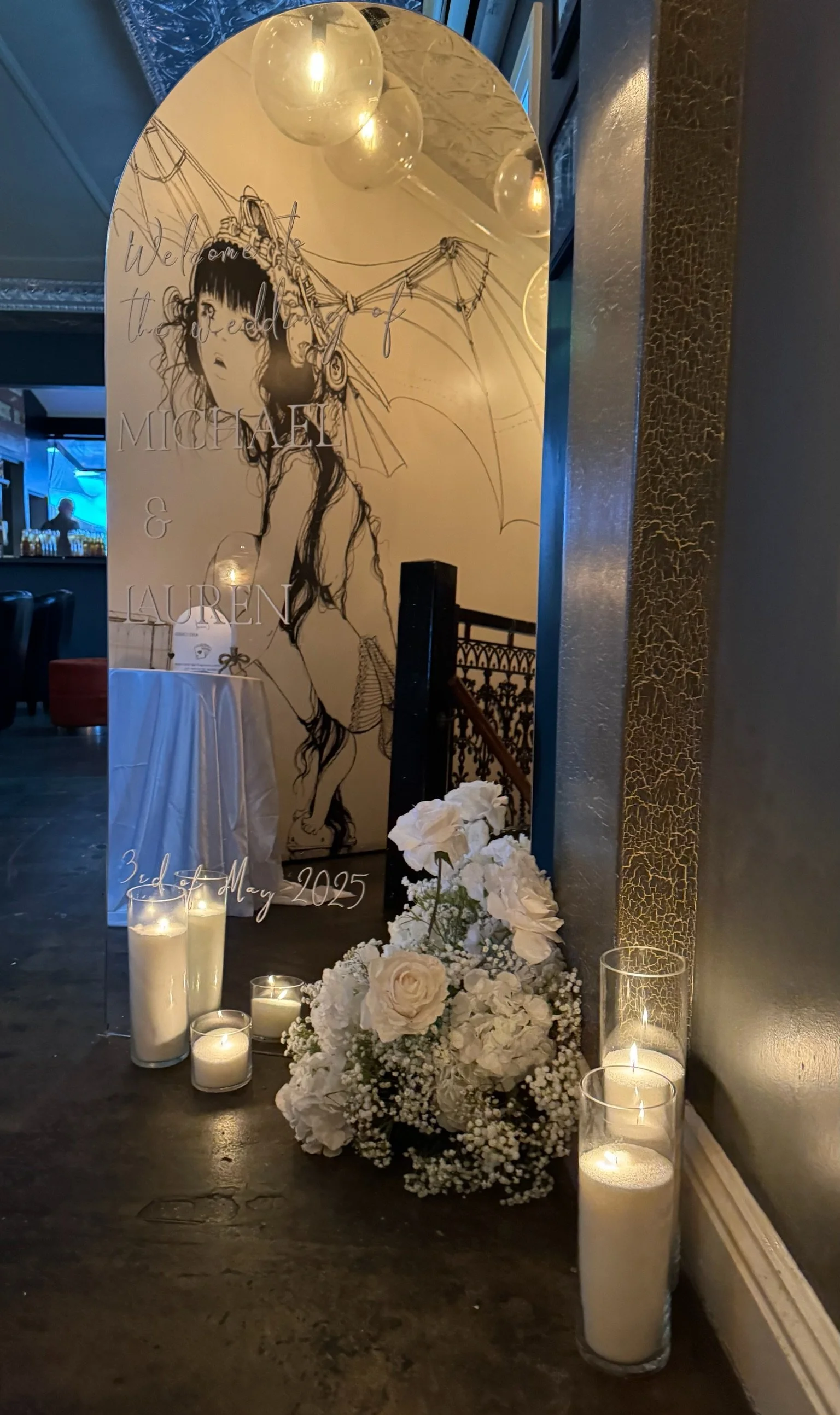 A wedding memorial with a mirror, candles, and a floral arrangement, featuring artwork of a girl with an umbrella, with details about the wedding date, May 3rd, 2025, and names Michael and Lauren.