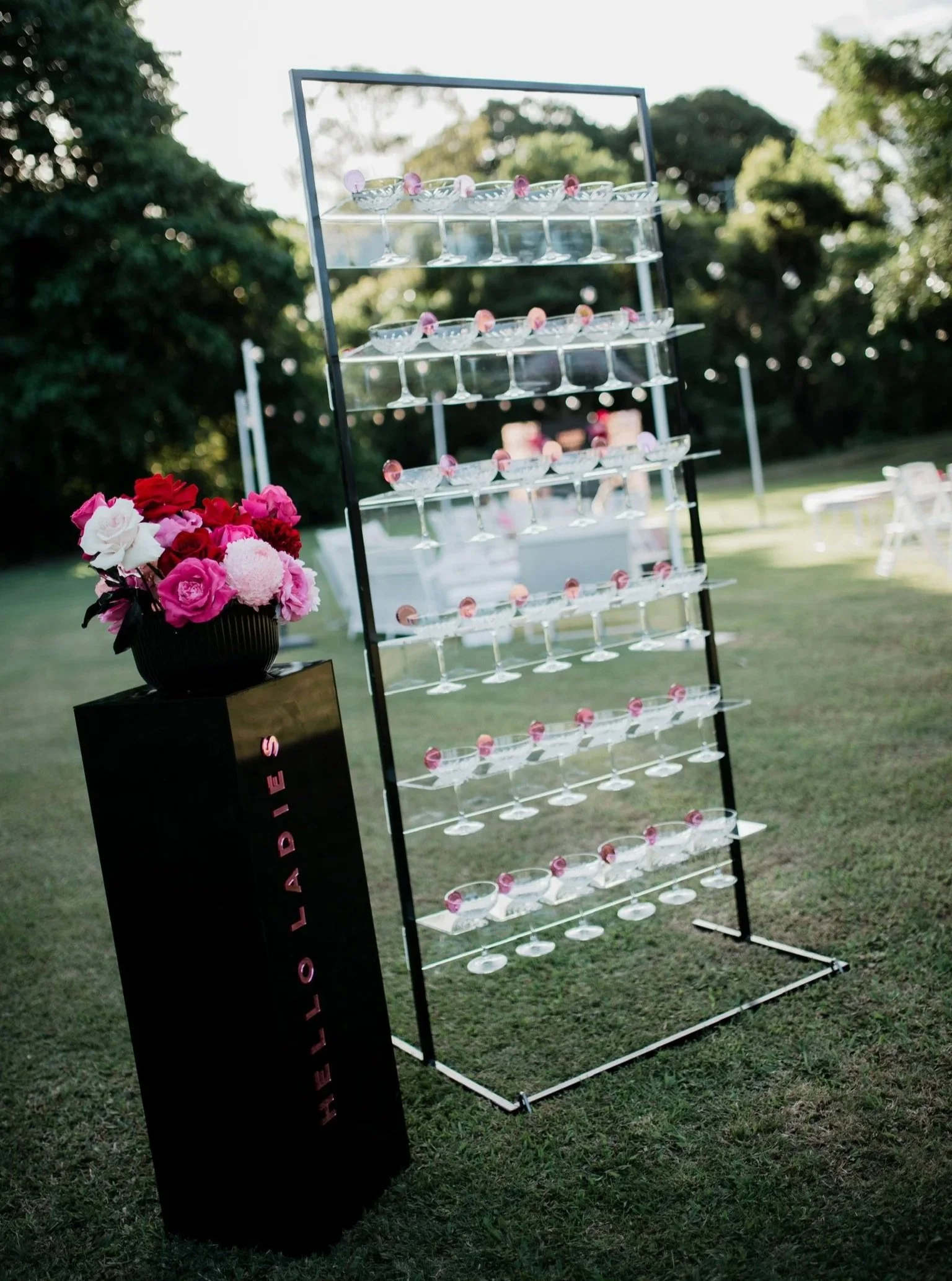 A display of empty cocktail glasses on a multi-tiered stand outdoors, with a black pedestal holding a bouquet of pink, red, and white flowers next to it, set in a grassy area with trees in the background.