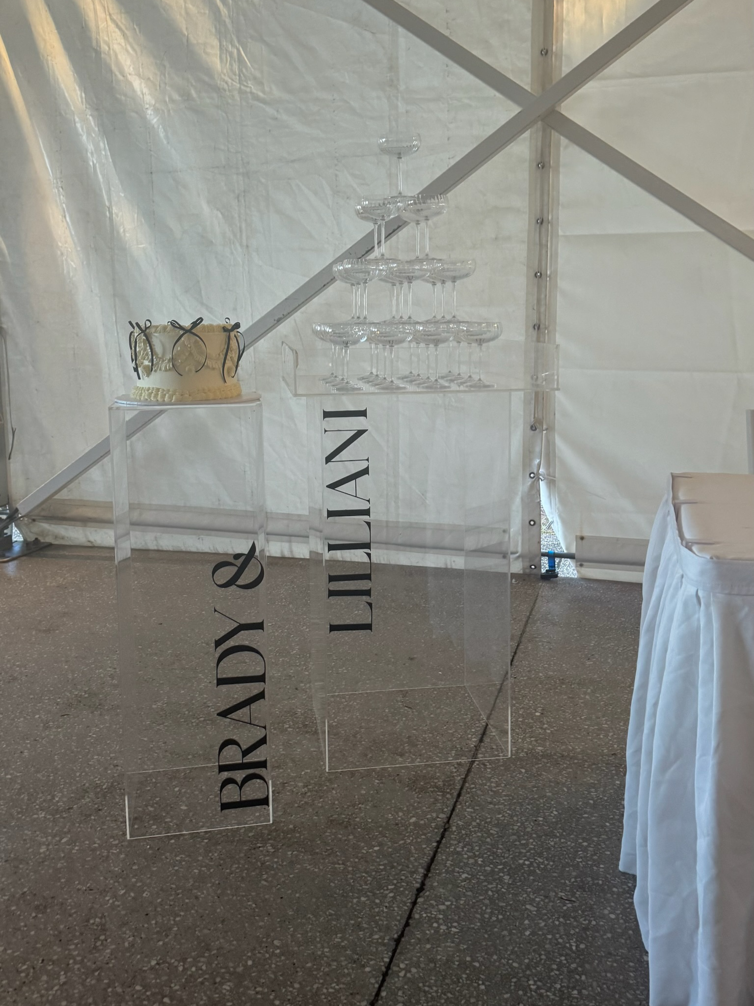 Cake and champagne glasses on transparent acrylic tables under a tent, with the sign reading "BRADY &amp; LELIANA" in black lettering.
