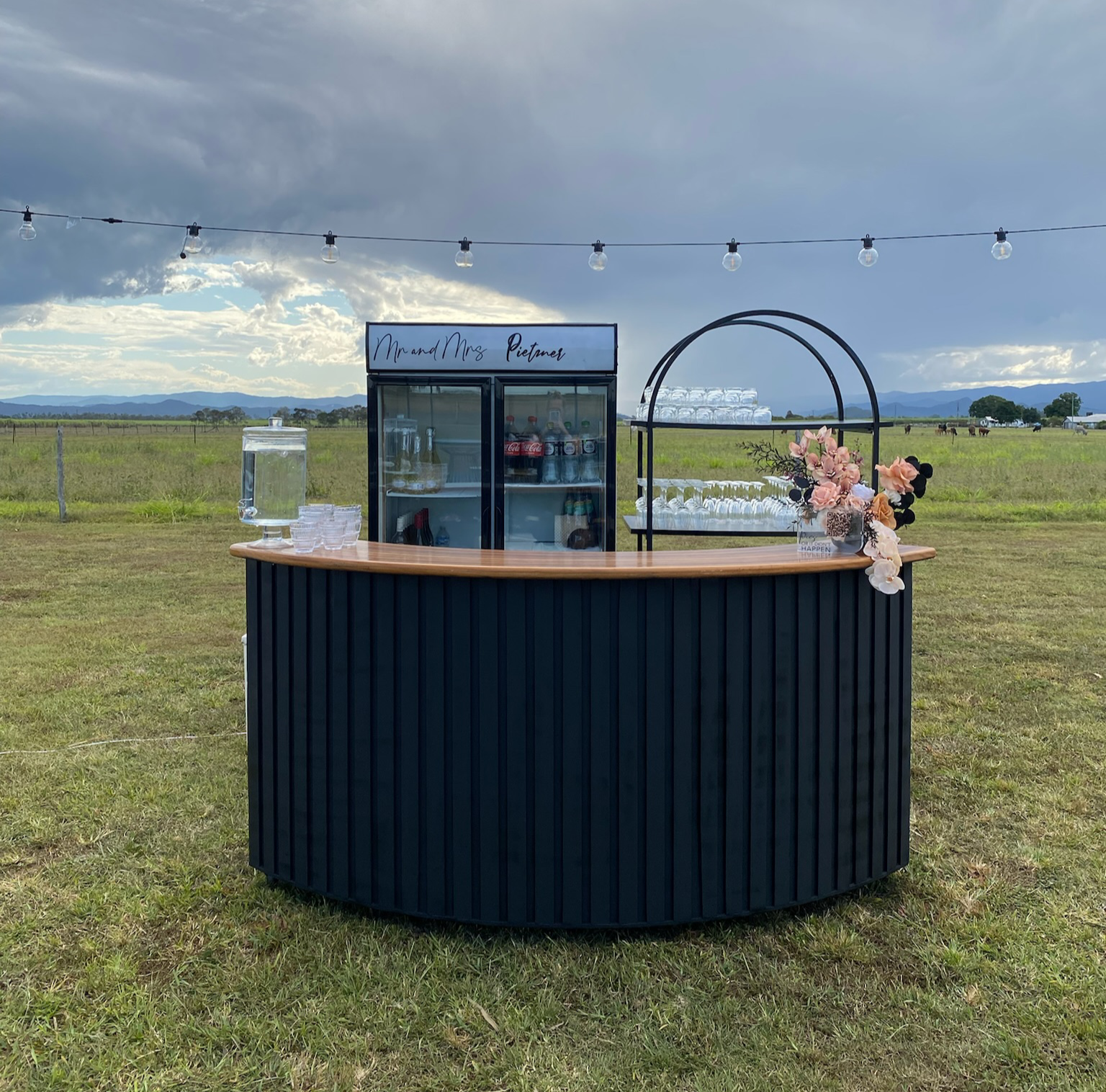 Outdoor beverage stand with black base and wooden top, decorated with pink and beige flowers, set in a grassy field with mountains in the background and string lights overhead.