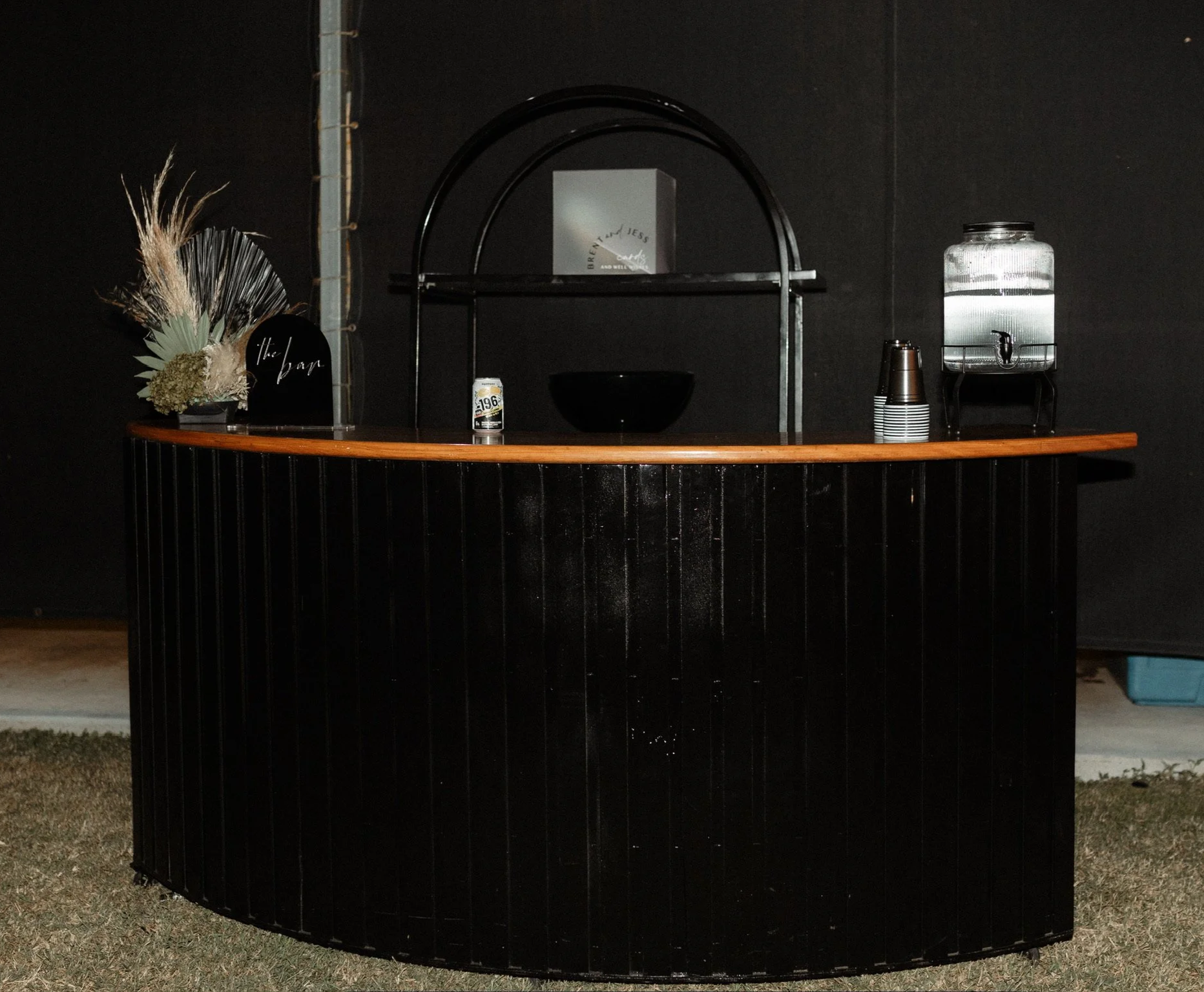 A black outdoor bar with a wooden top, decorated with a floral arrangement, black bowl, drink can, stack of cups, and a water dispenser, set against a black wall.