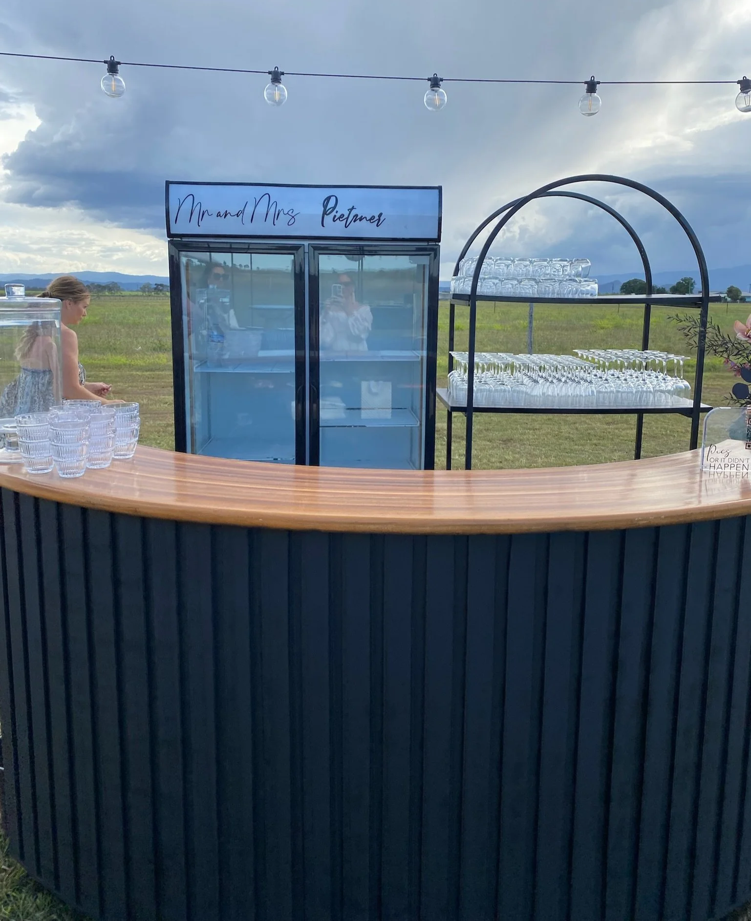 Outdoor bar setup with glasses, a glass-door refrigerator, and a glass shelf, with a cloudy sky and fields in the background.
