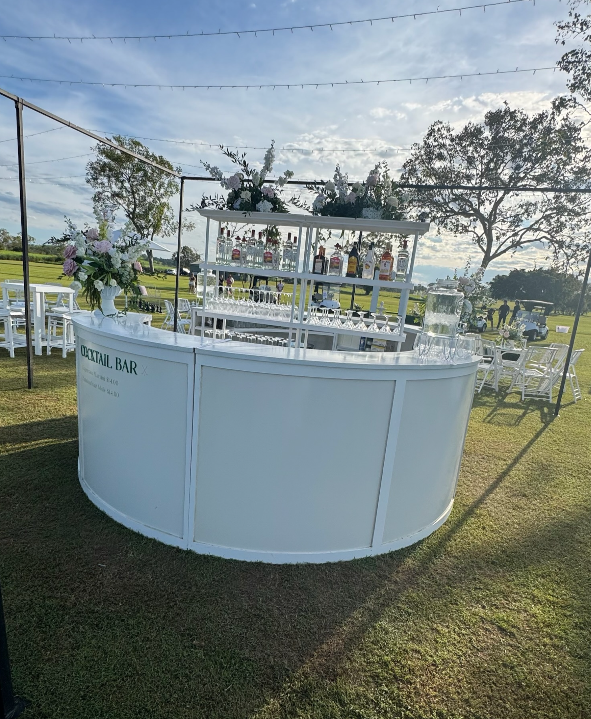 White outdoor cocktail bar with floral arrangements and various bottles of alcohol on a tiered shelf, set up on grass with tables and chairs in the background, under a partly cloudy sky.