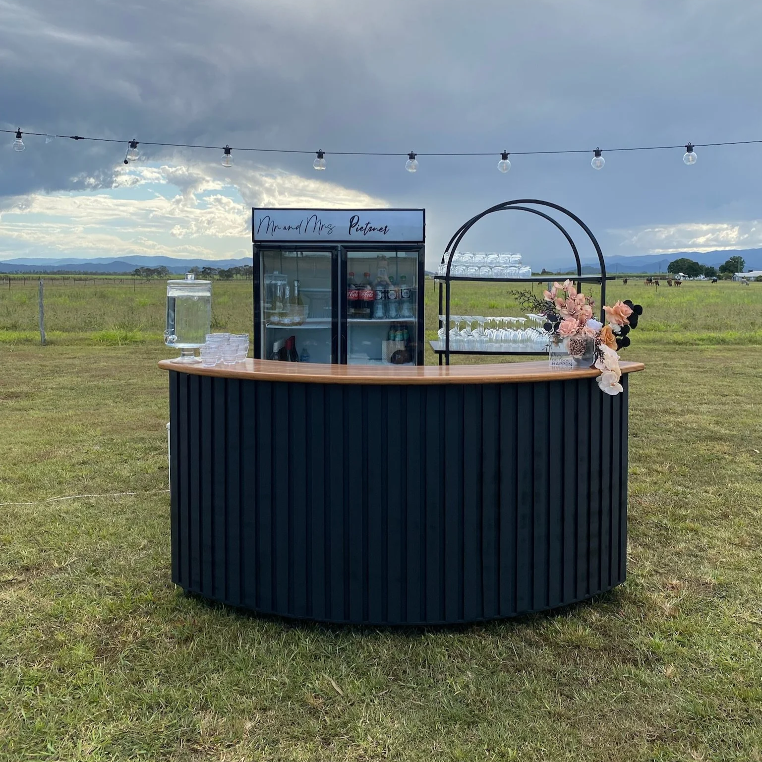 Outdoor bar setup with black curved counter, flower arrangement, drink glasses, water dispenser, and beverage fridge in a grassy field with cloudy sky and mountains in the background.