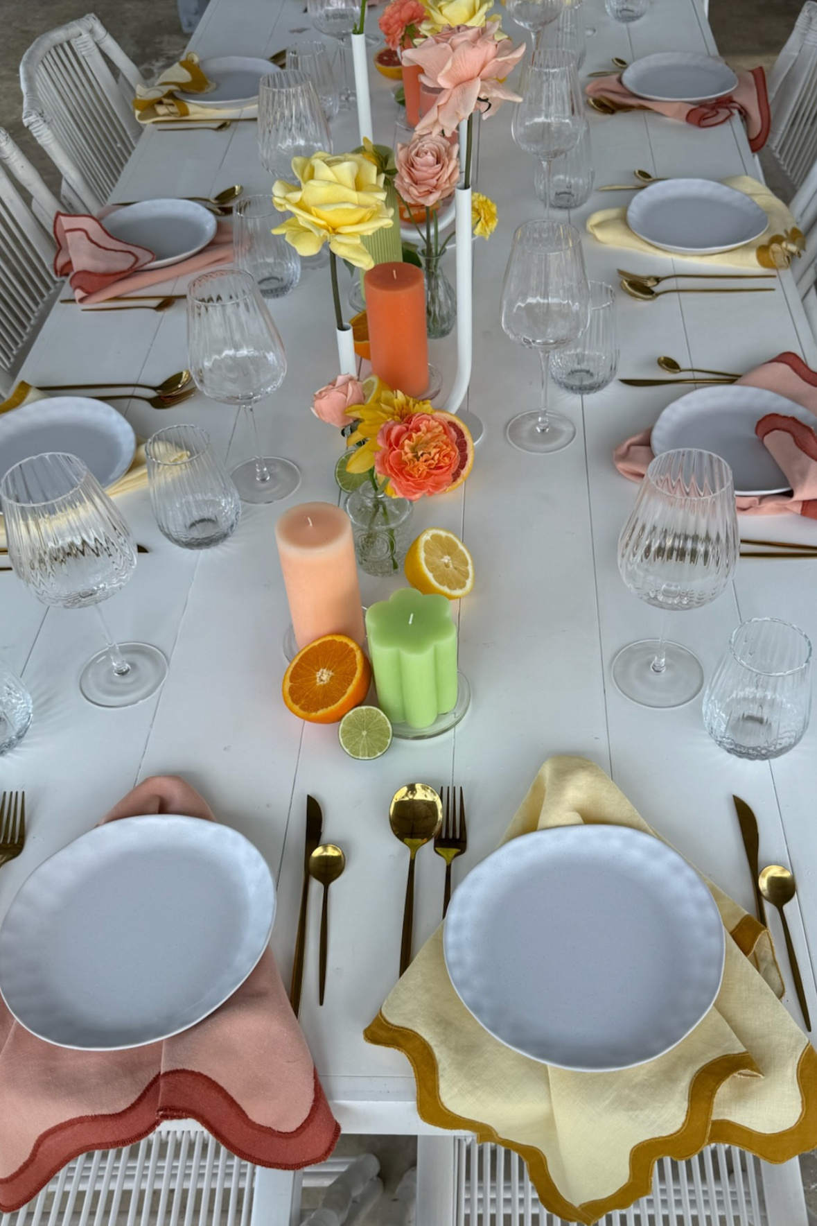 A table set for a meal with white plates, gold utensils, pastel-colored napkins, and glassware. The center features candles, flowers, and slices of citrus fruits.