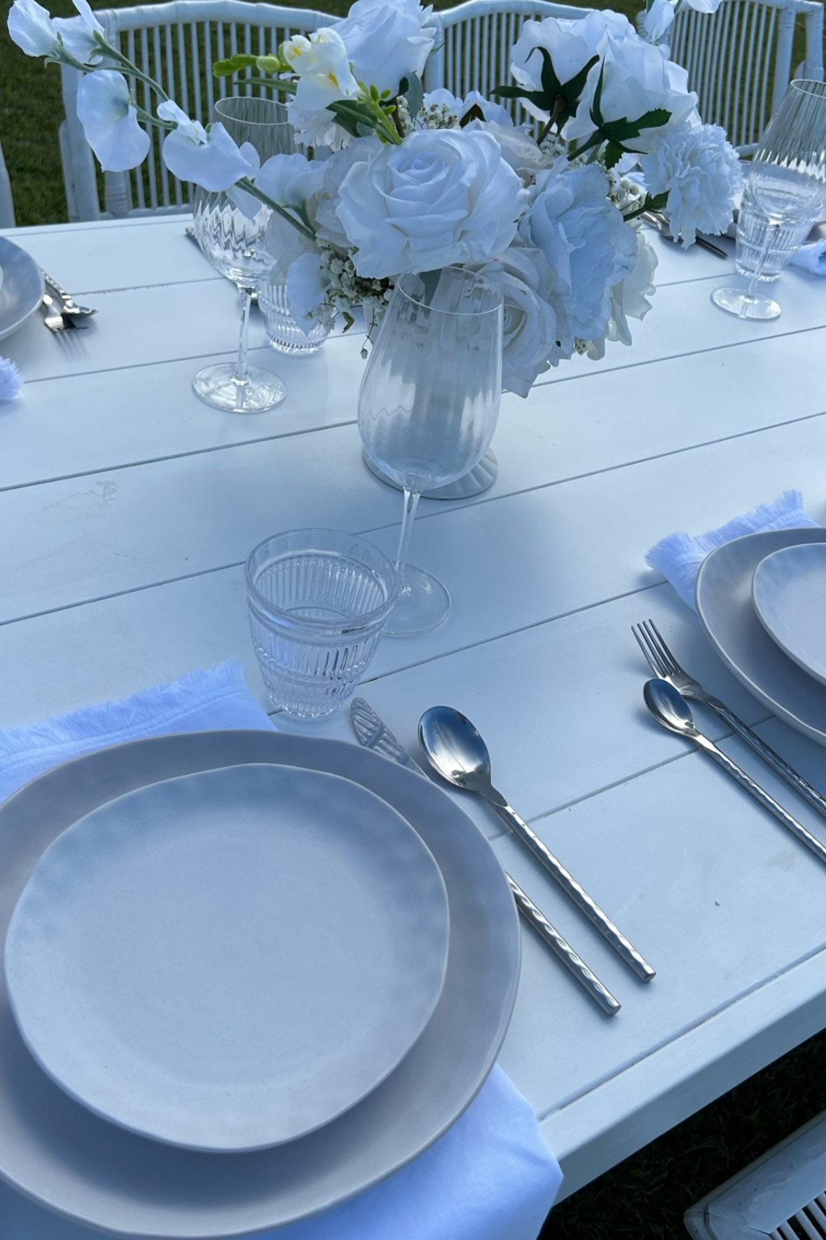 A white outdoor dining table with a floral centerpiece of white flowers in a clear glass vase, surrounded by clear glassware and white plates with silver cutlery, set for a formal meal.