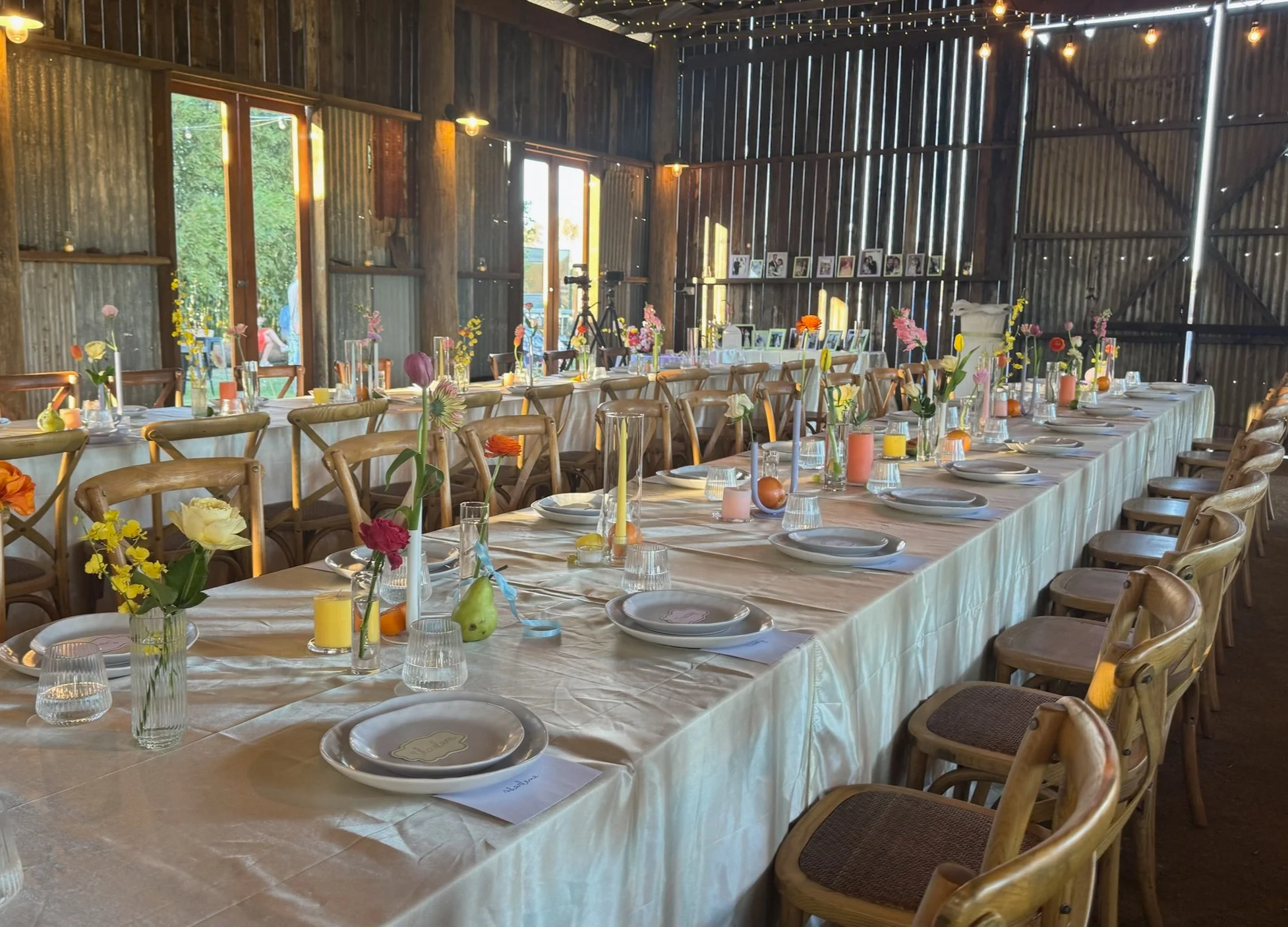 Long table set for a celebration or event inside a rustic barn, decorated with colorful flower centerpieces in vases, candles, plates, glasses, and napkins, with wooden chairs arranged along the sides.