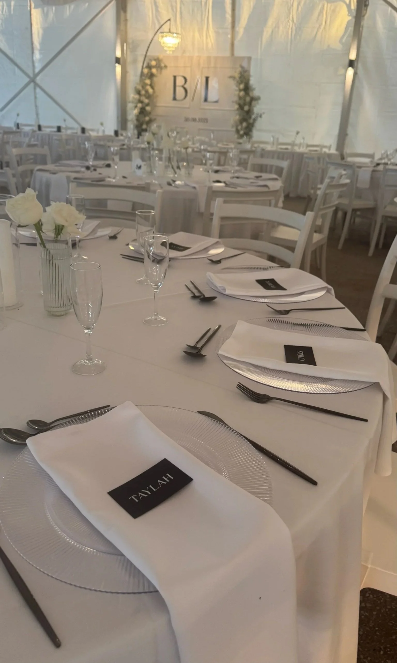 Elegant wedding reception table setup with white tablecloths, glassware, silverware, white napkins, and place cards with names Talayah and Oliva, inside a decorated tent with a sign displaying initials 'B' and 'L' and the date 30.10.2023, surrounded 