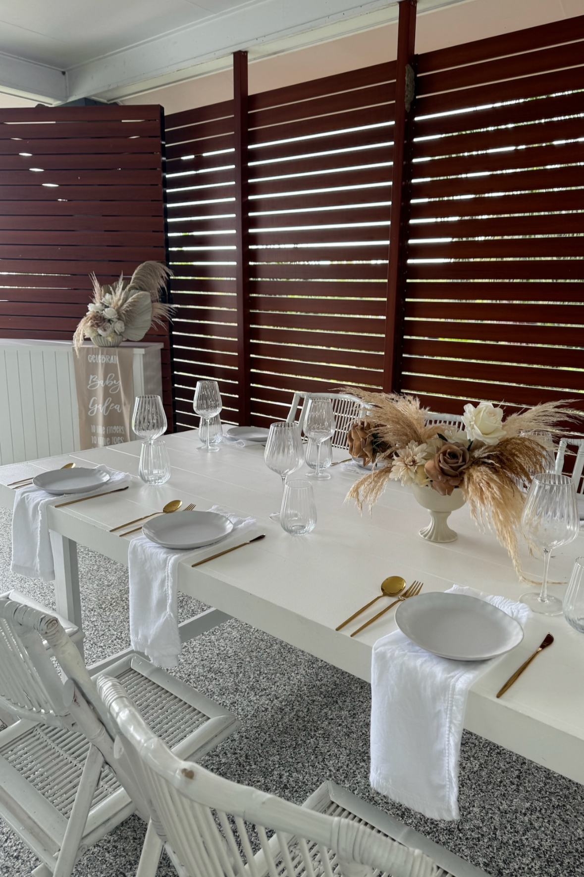 A decorated dining table set for a celebration with white plates, gold utensils, empty wine glasses, and a floral centerpiece featuring white and brown flowers with pampas grass. The table is on a speckled gray floor, with a wooden privacy fence in t
