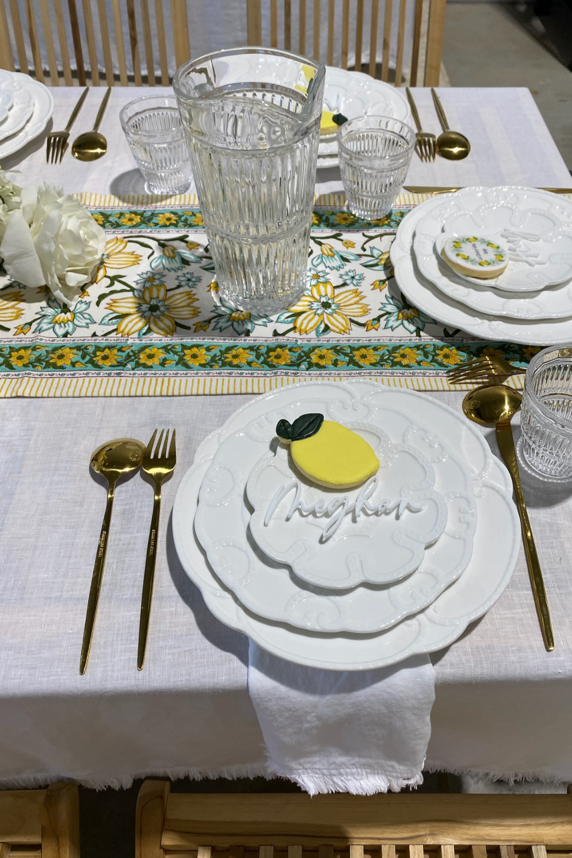 A decorated place setting for a meal with white plates, gold utensils, clear glasses, and a centerpiece with white flowers. There is a decorative cookie shaped like a lemon with green leaves and the word "Cheekar" written on it placed on the foregrou