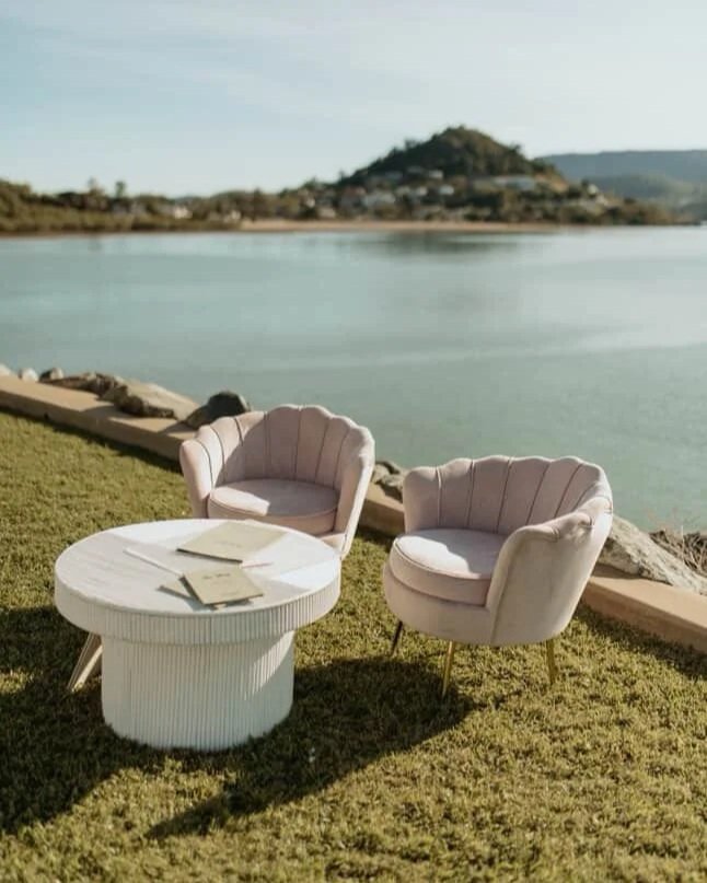 Two pink velvet armchairs and a white ruffle coffee table on a grassy area beside a large body of water, with a mountain and hilly landscape in the background, under a cloudy sky.