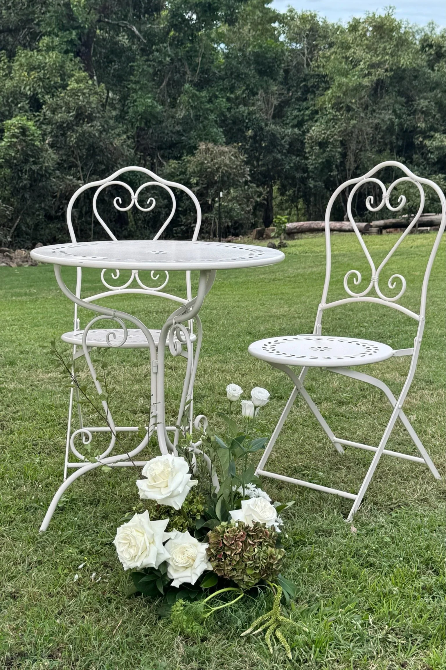 A white wrought iron outdoor table and two matching chairs are set on a grassy lawn with flowers at the base, and a trees background.