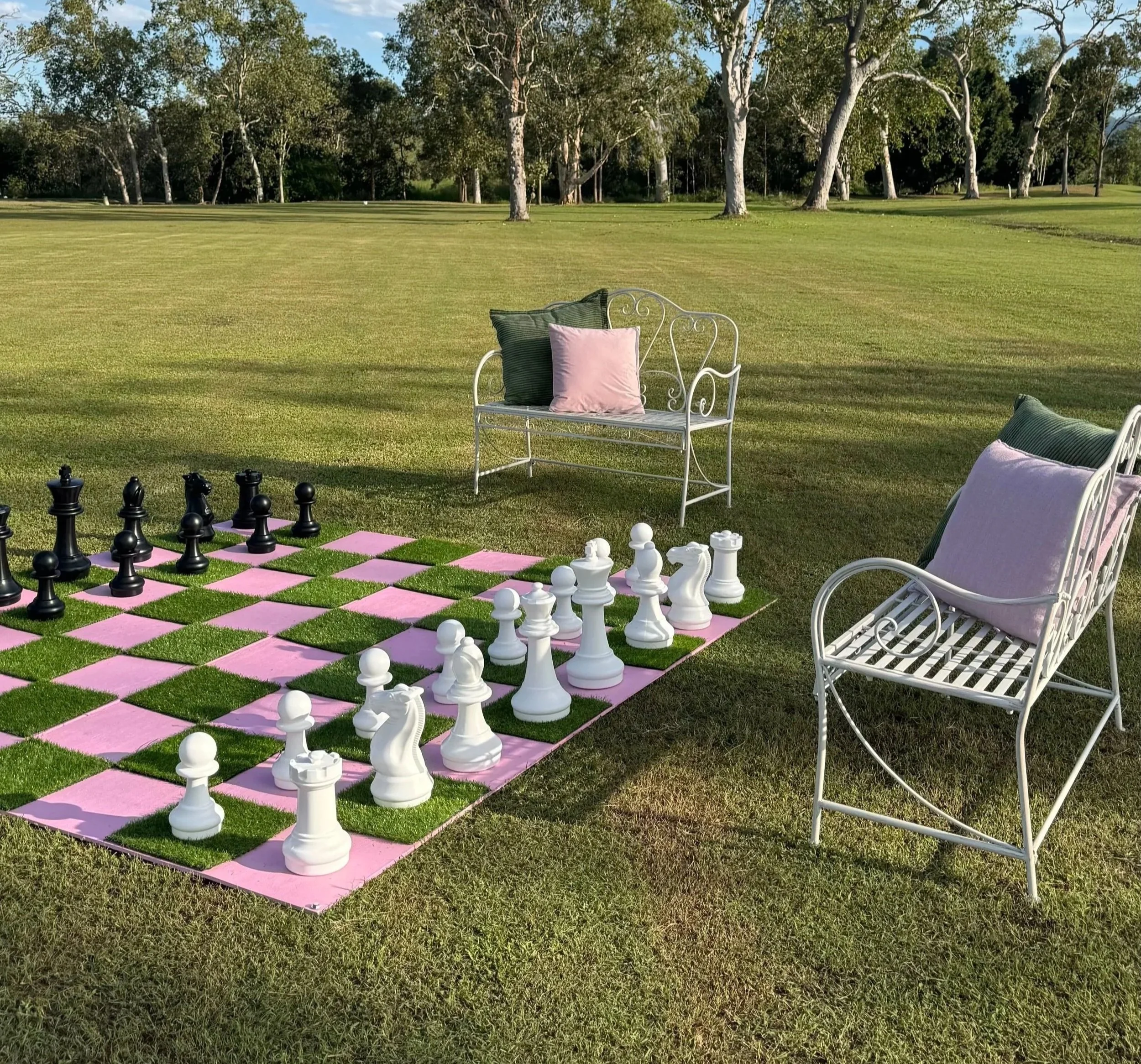 A backyard with a pink and green chessboard on the grass, two white metal benches with pink and green cushions, and large trees in the background.