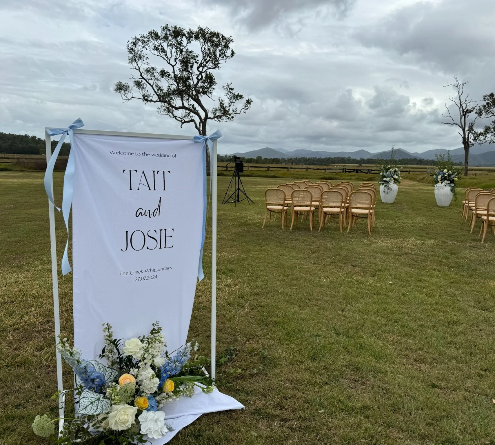 Outdoor wedding setup with a white sign displaying 'Welcome to the wedding of Tait and Josie, The Creek Whitsundays, 27.07.2024', decorated with flowers. There are rows of wooden chairs and white floral arrangements in large vases on a grassy field w
