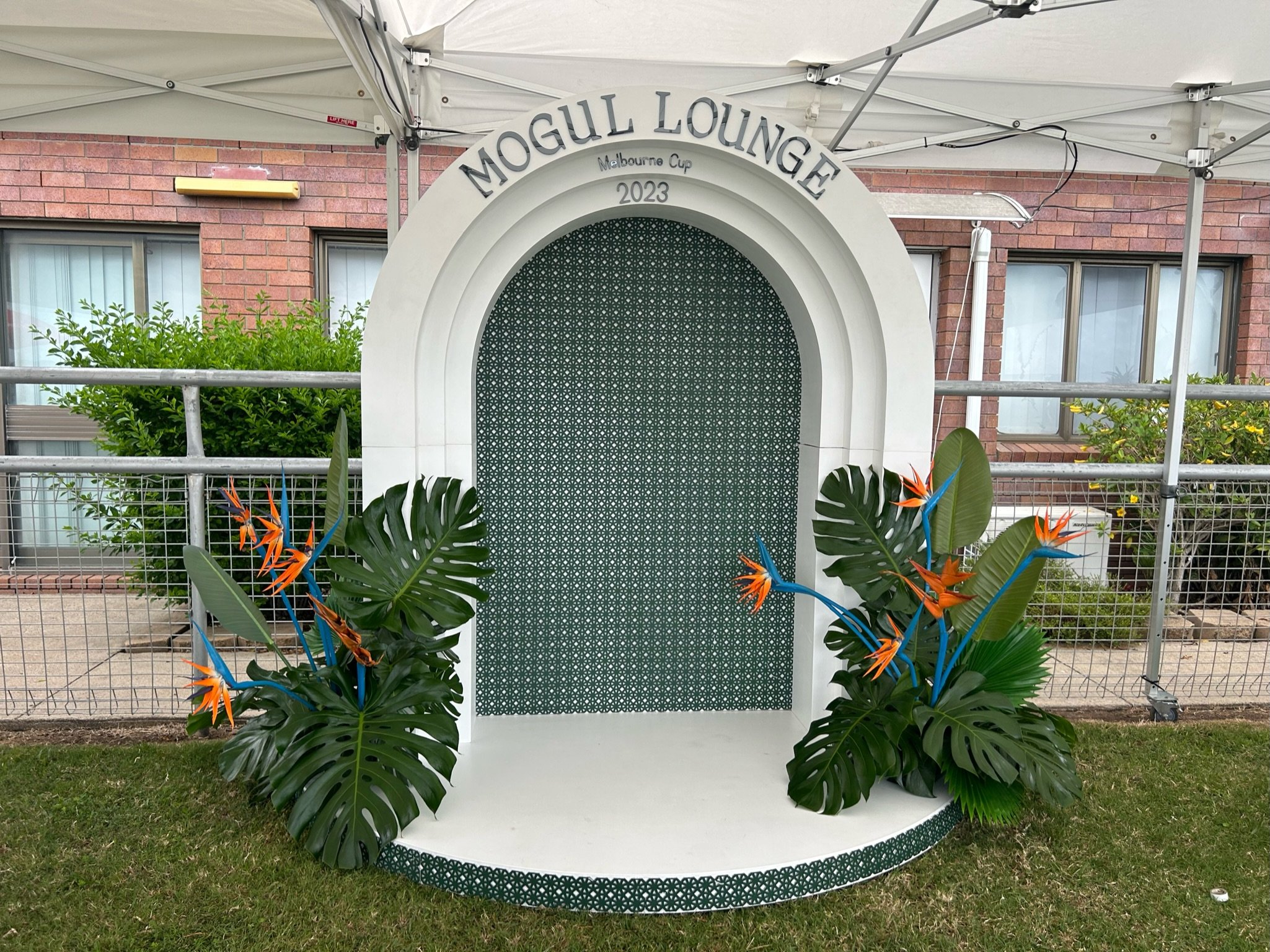 Decorative arch with the text 'Mogul Lounge' at the top, surrounded by tropical plants with orange and blue flowers, set against a fenced outdoor area with a brick building in the background.