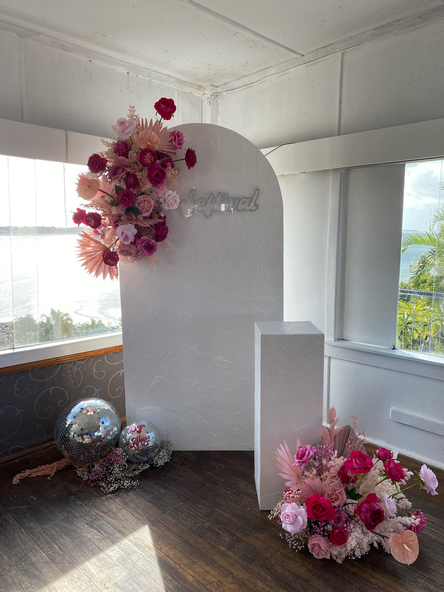 Decorative photo backdrop with pink, purple, and peach floral arrangements, two disco balls, and a white sign that says 'Love' in cursive, set in a room with large windows and wooden floors.