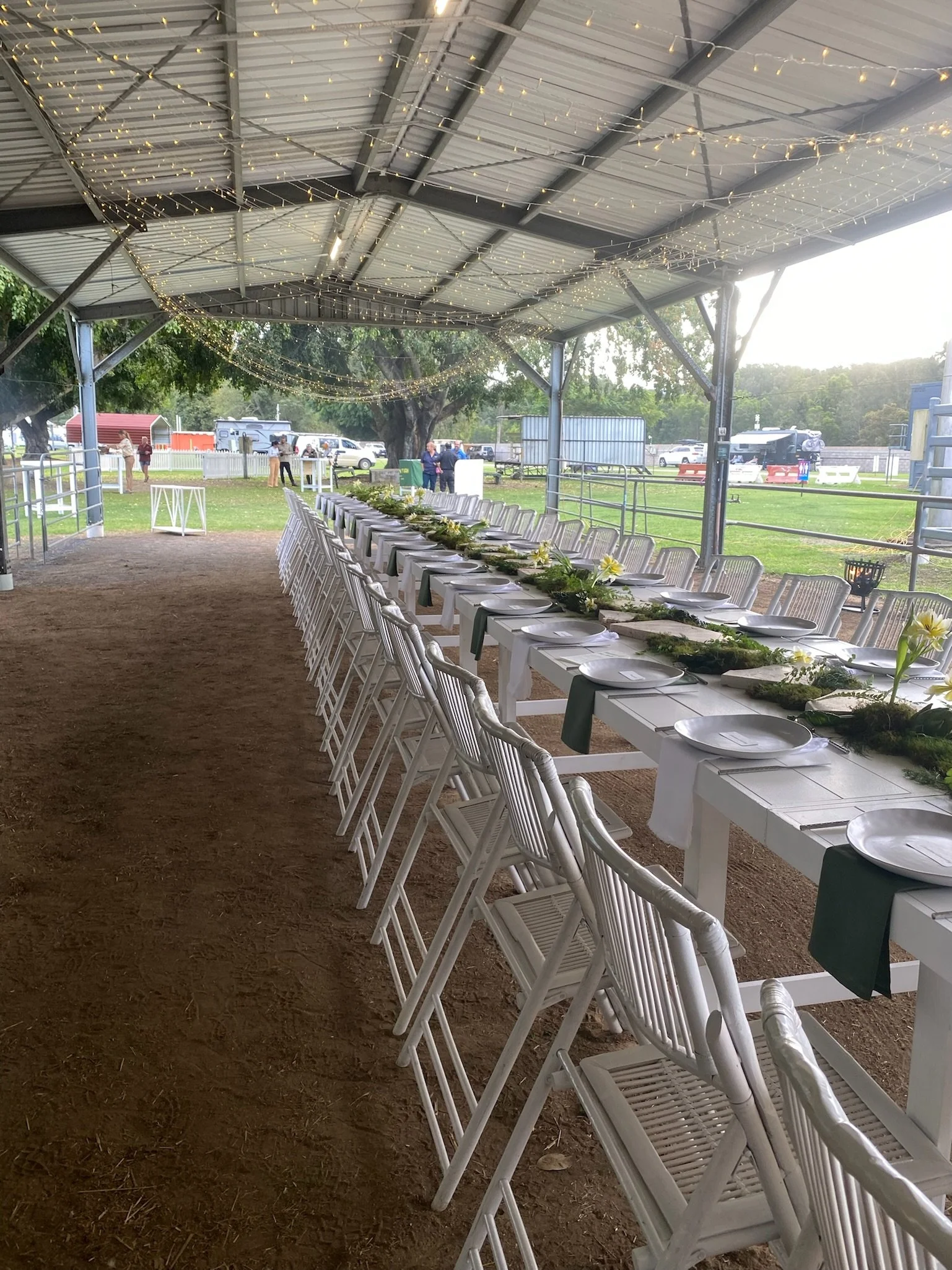Long table set under a decorated outdoor pavilion with string lights, white chairs, and tableware, prepared for a gathering.