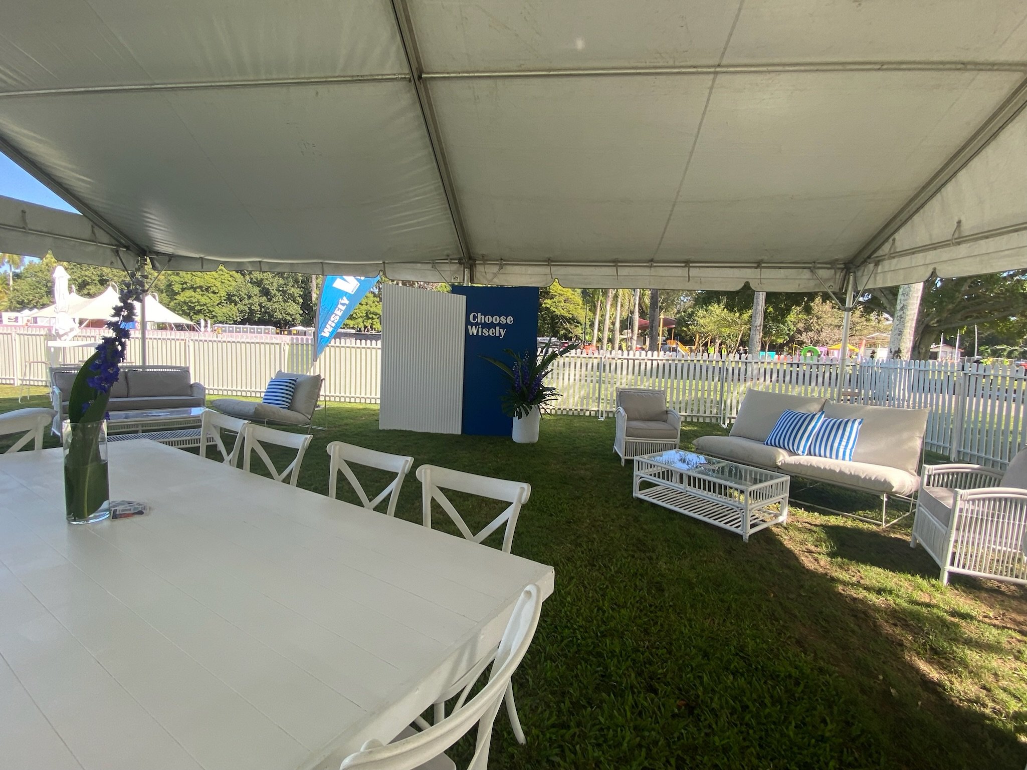 Empty outdoor event tent with white tables and chairs, white couches with blue striped pillows, and a potted plant inside, set up on green grass with a white picket fence and trees in the background.