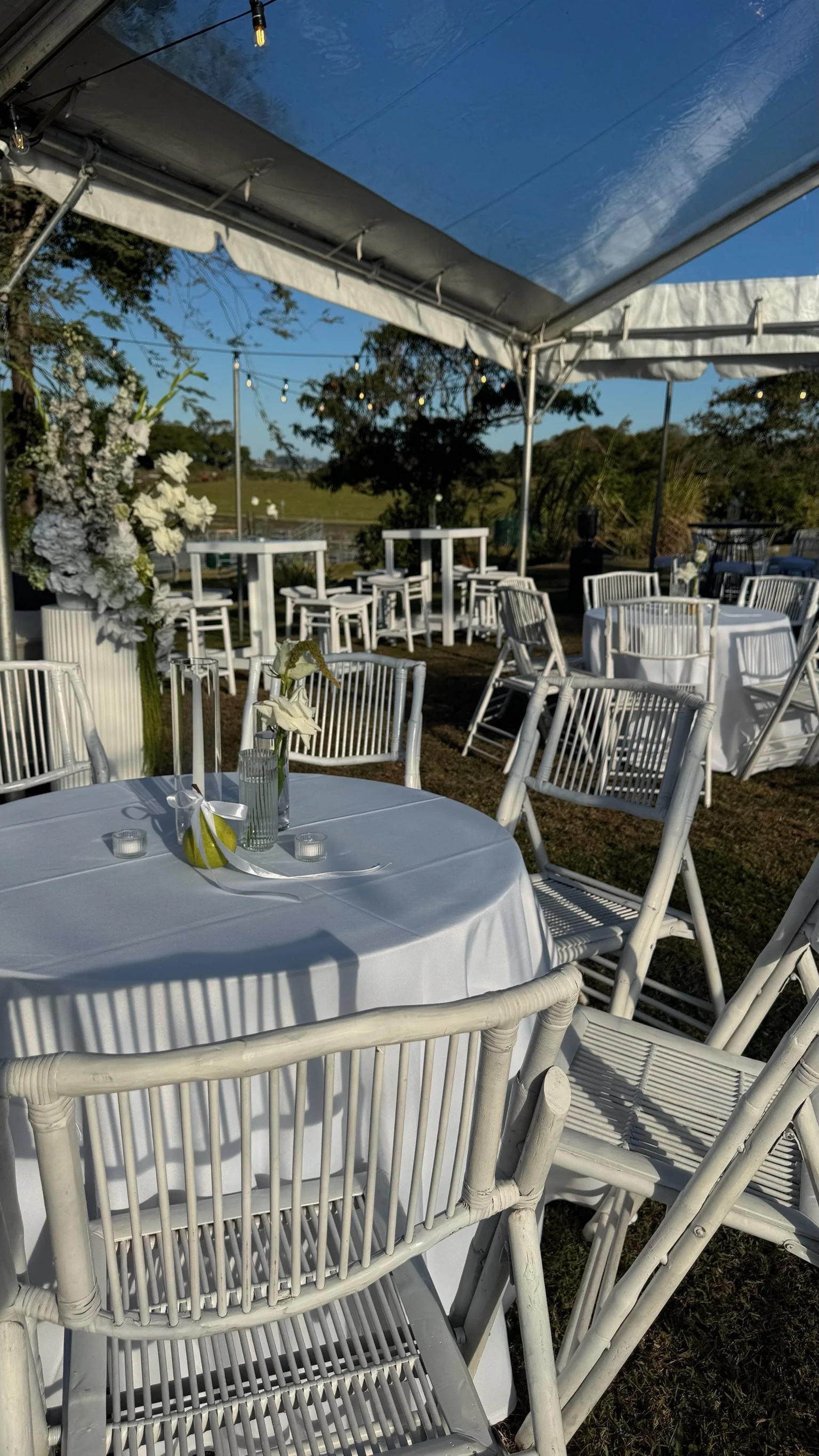Outdoor wedding reception setup with round tables covered in white tablecloths, white chairs, floral centerpieces, and string lights under a blue canopy.