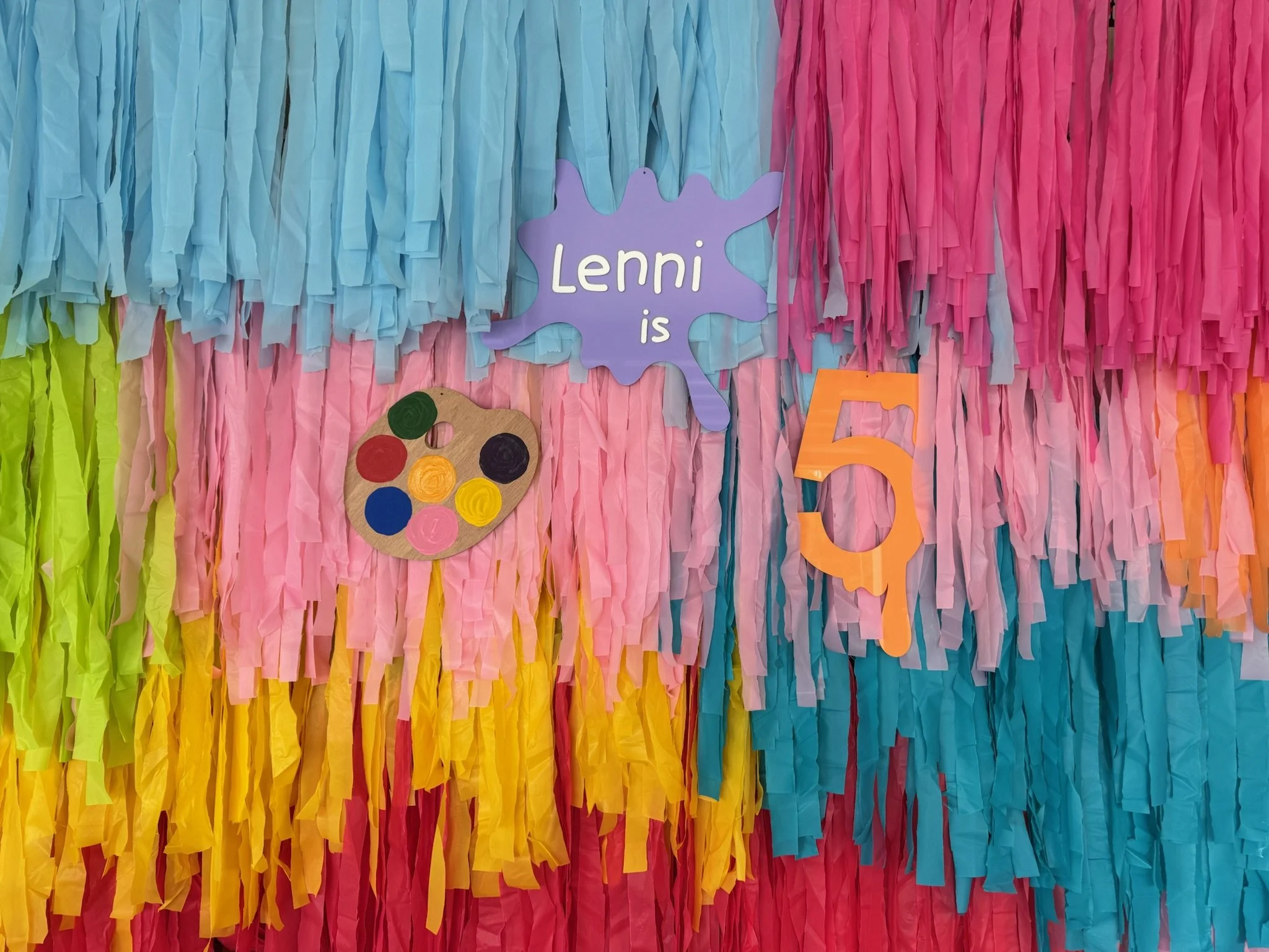 Colorful tissue paper backdrop with a sign that reads "Lenni is 5," a painter's palette decoration, and a large orange number 5 cutout.