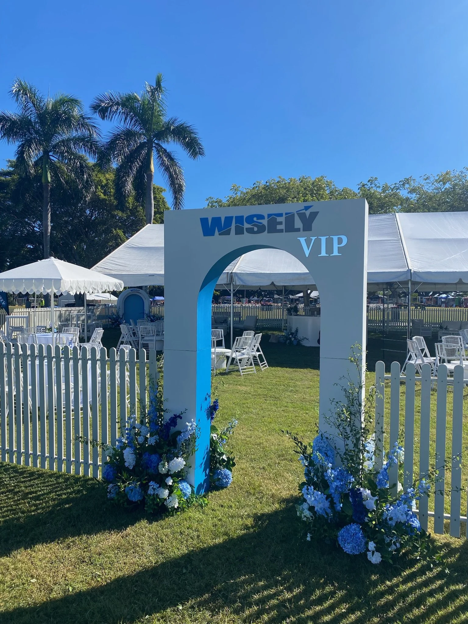 Entrance to an event with a white and blue sign reading 'WISELY VIP' surrounded by flower arrangements, white fencing, and a white canopy tent in the background under a clear blue sky.