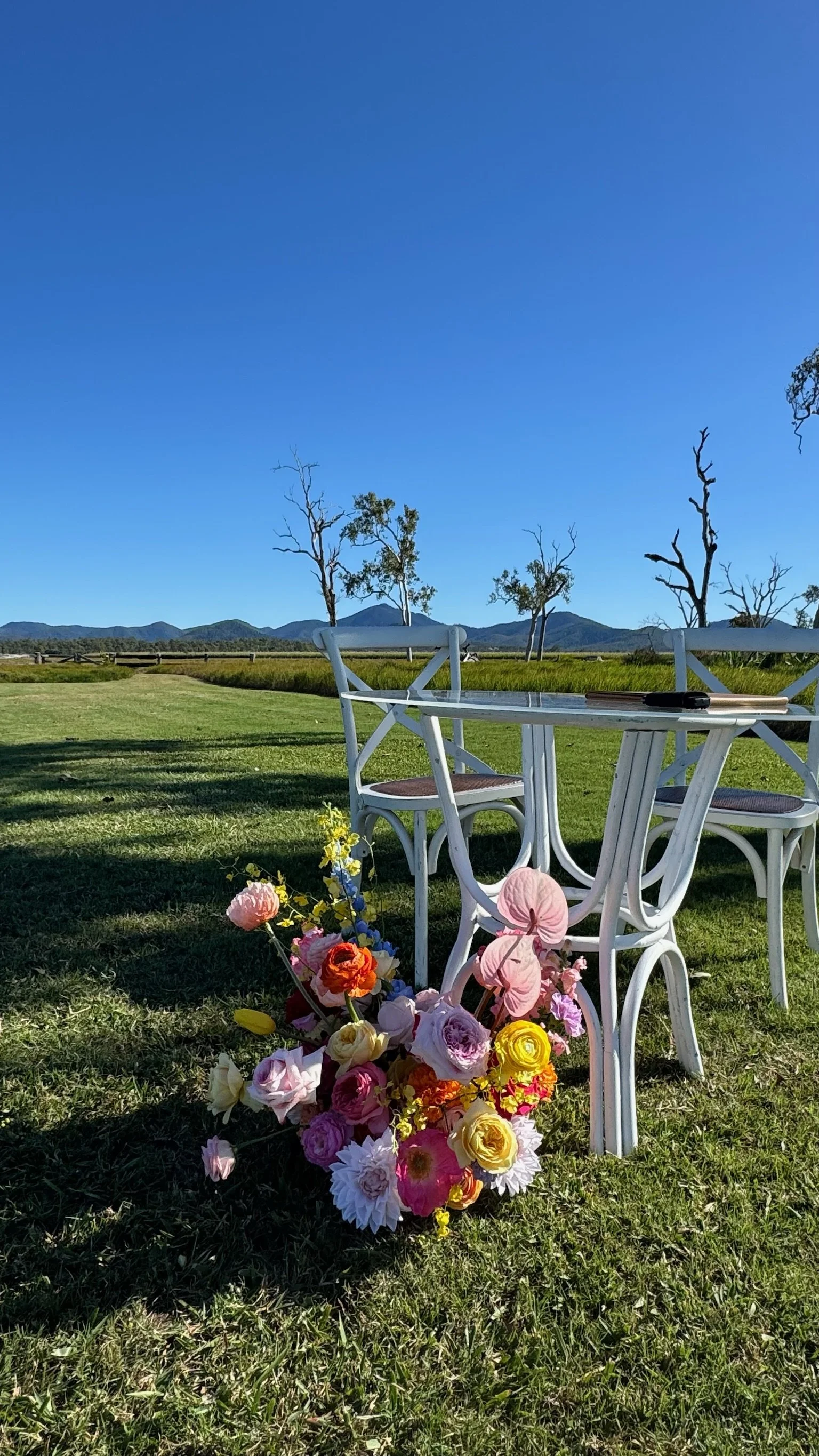 A white outdoor table and chairs set on a grassy field with a bouquet of colorful flowers in front, under a clear blue sky with a few trees and distant mountains in the background.