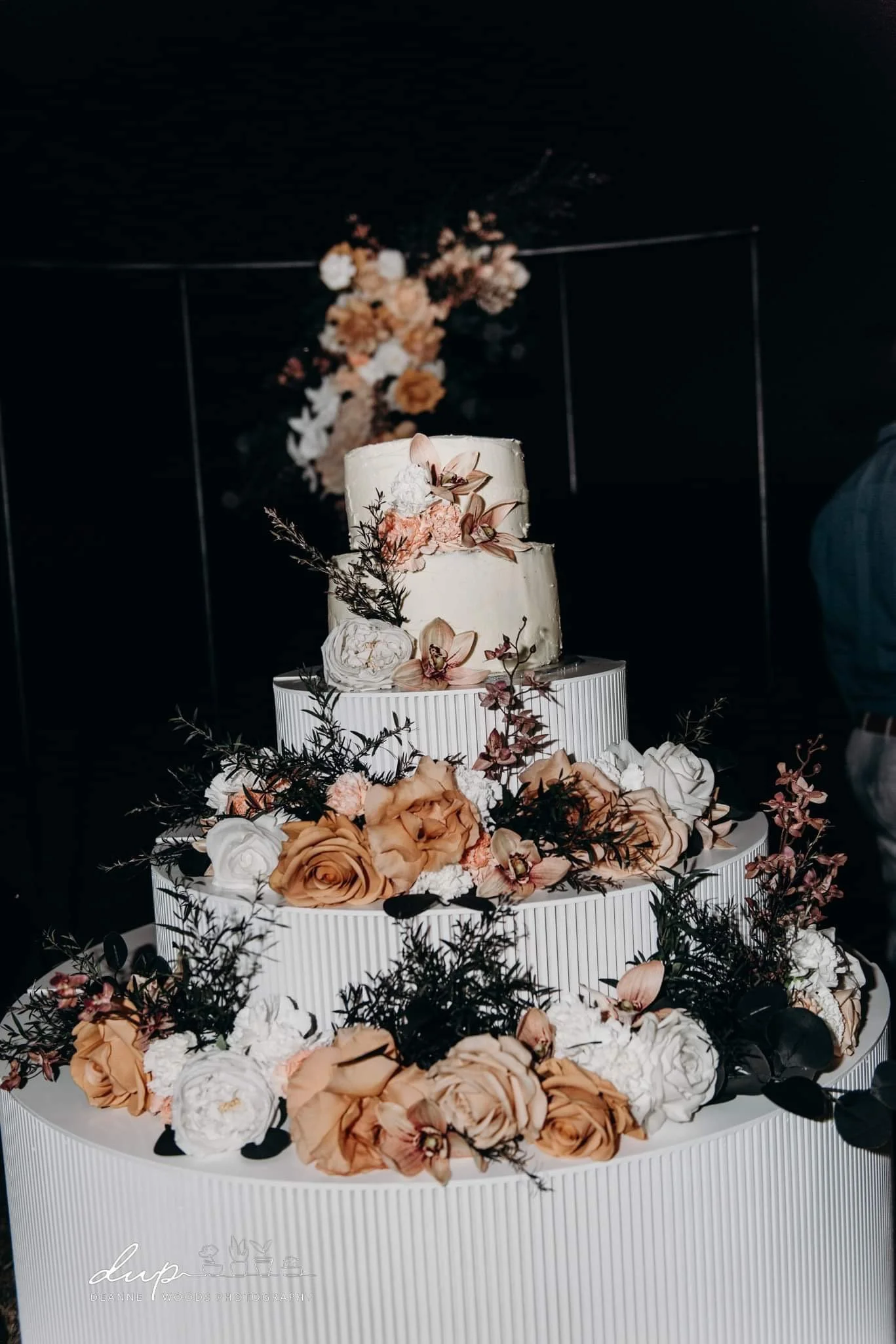 Three-tier wedding cake decorated with peach and white roses, orchids, and greenery, placed on a white textured platform with a floral backdrop in the background.