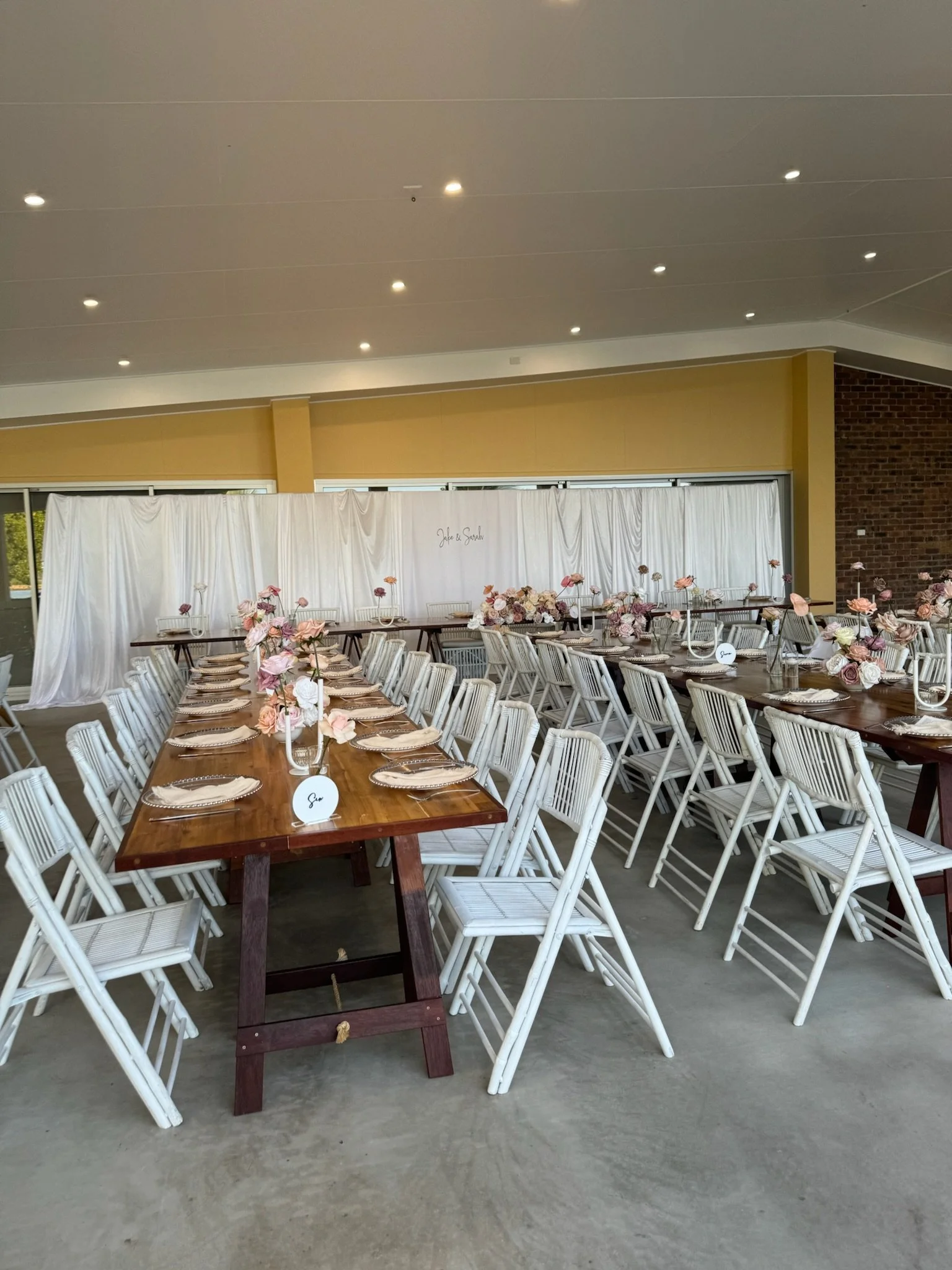 Wedding reception area with long wooden tables decorated with pink and white floral centerpieces, white chairs, and a white draped backdrop with handwritten names 'Jade & Sarah'.