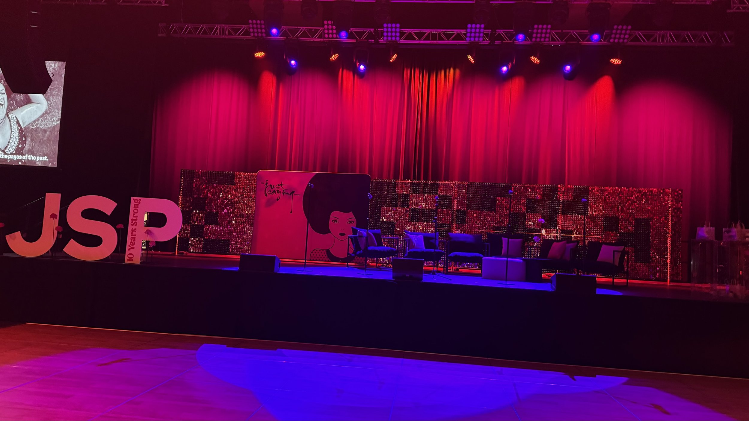 Stage with red curtains, seating area with chairs and sofas, large sign reading "JSP 10 Years Strong," and a colorful backdrop featuring a stylized illustration of a woman with an Afro hairstyle, illuminated by stage lights.