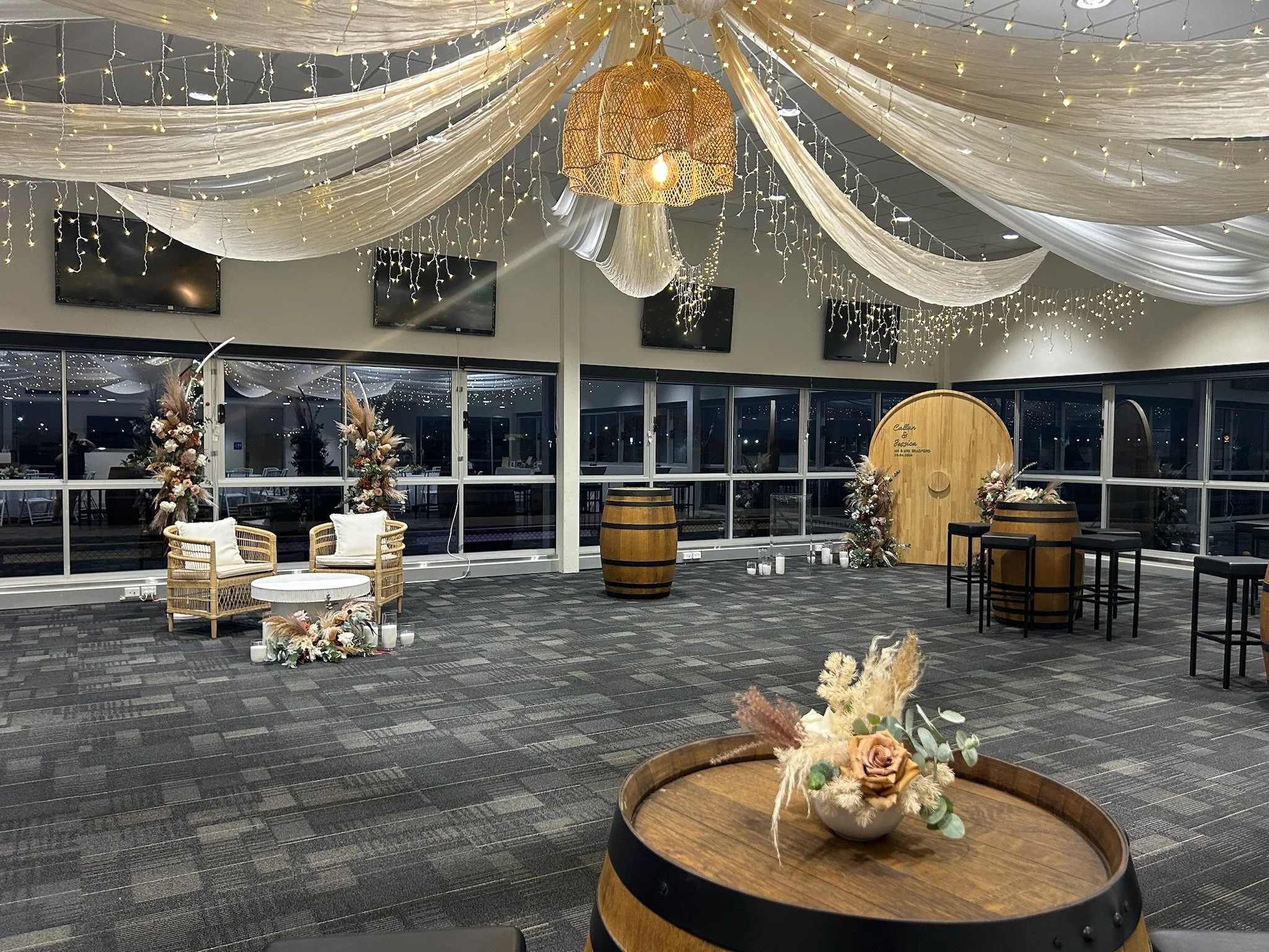 Decorative event space with draped fabric, fairy lights, and floral arrangements on tables and barrels, ready for a celebration or wedding.