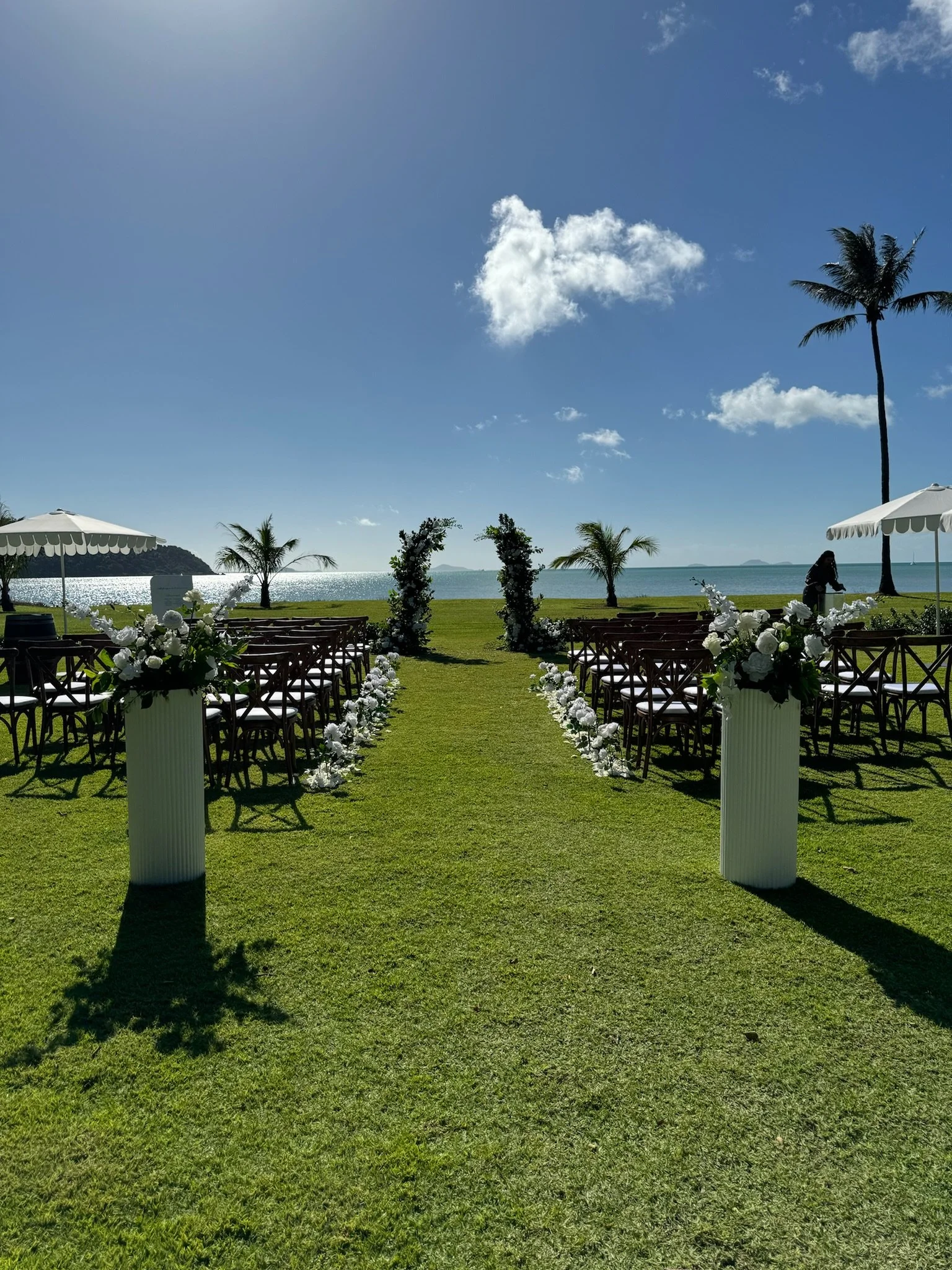 Outdoor wedding setup with chairs facing an arch decorated with flowers, on a grassy area by the ocean with palm trees and umbrellas.
