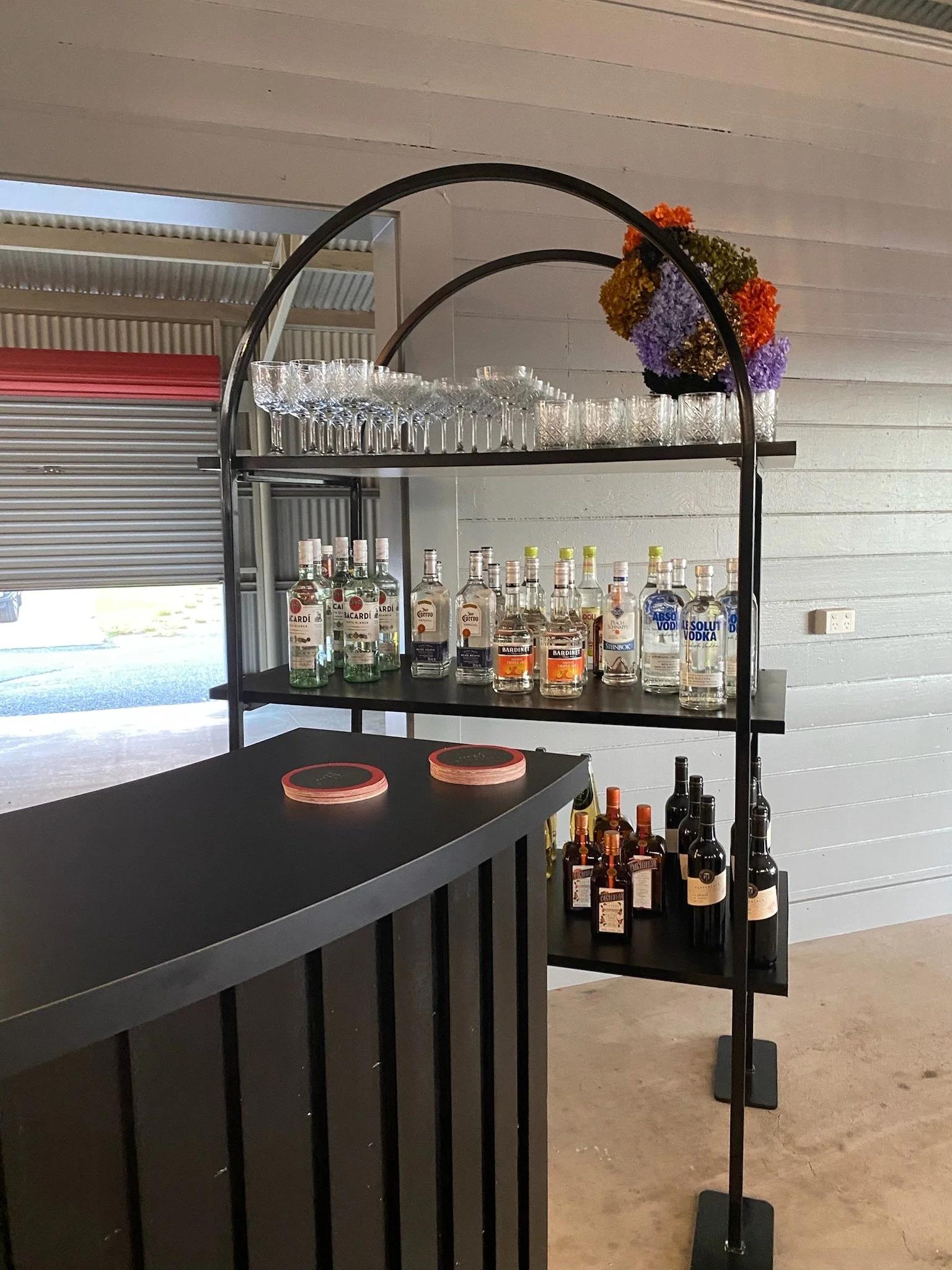 A black bar cart with three shelves holding glassware, bottles of liquor, and colorful flowers at a bar or event setting.