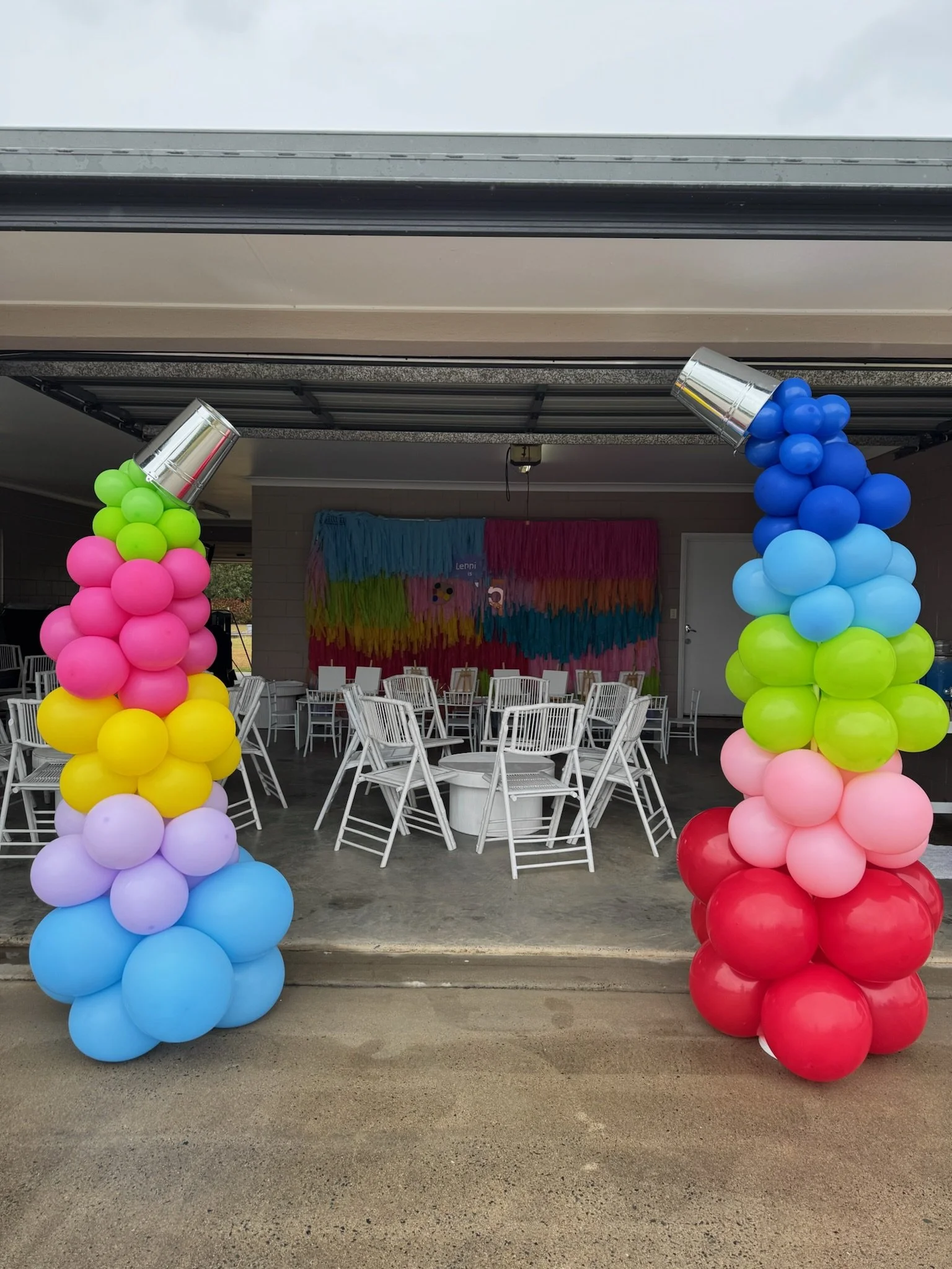 Colorful balloon arch doorway with pink, yellow, purple, blue, green, and red balloons, leading into decorated party space with tables, chairs, and rainbow-themed backdrop.