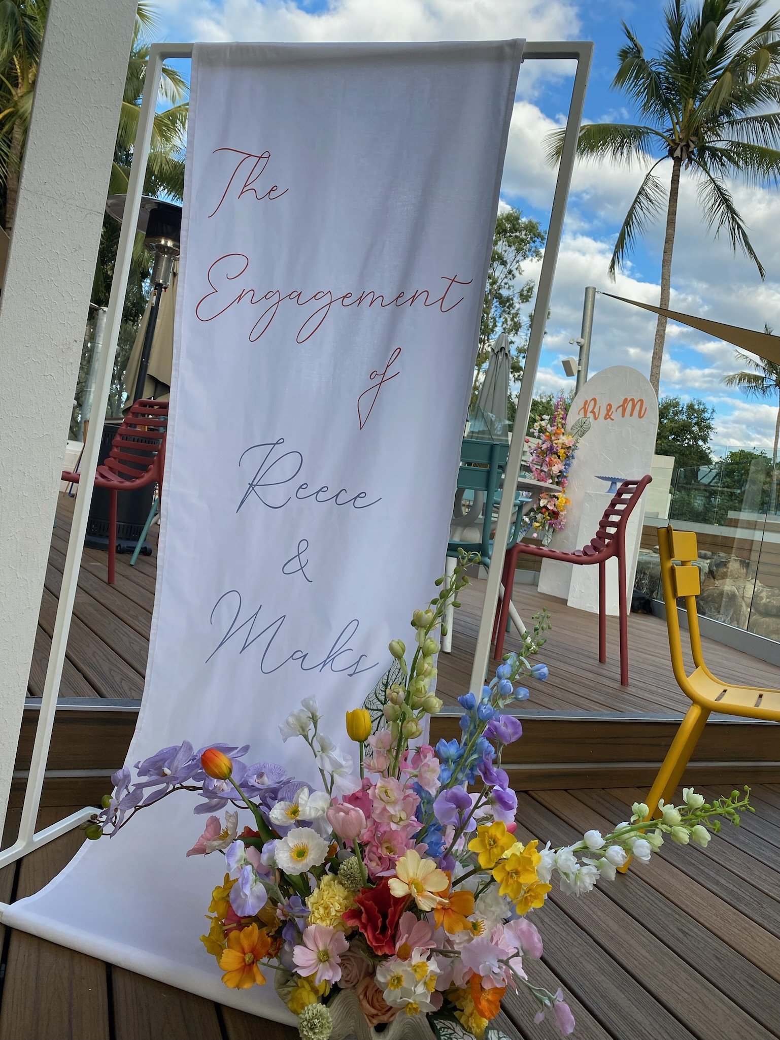 A large white banner with elegant cursive text reading 'The Engagement of Reece & Micks' on a metal frame at an outdoor venue, decorated with a colorful bouquet of flowers at the base, featuring pink, purple, white, yellow, and orange blooms, on a wo