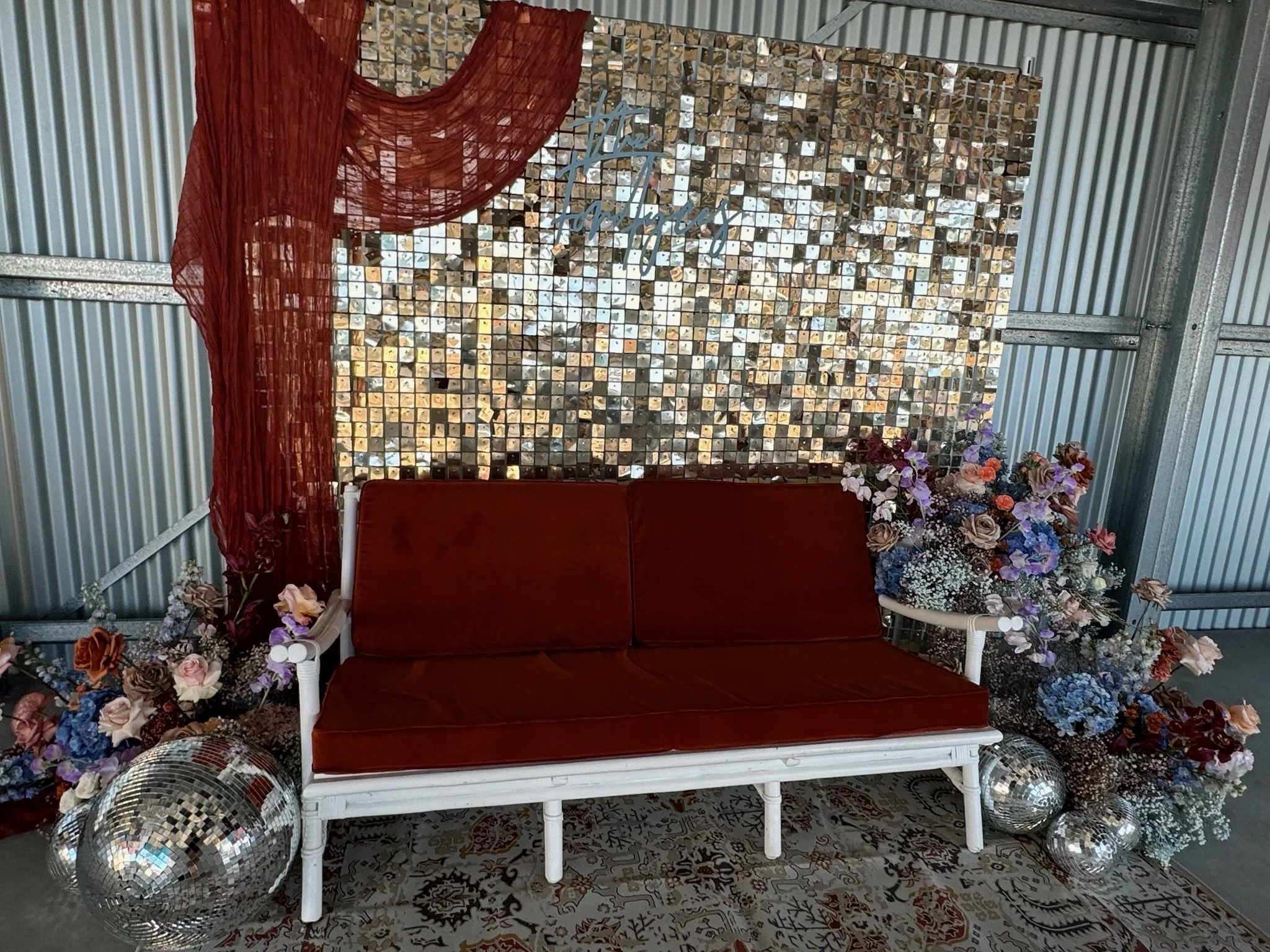 A vintage-style white bench with red cushions is placed in front of a decorated backdrop featuring a mosaic of small mirror tiles, a sheer red drape on the left, and large floral arrangements with purple, white, and pink flowers on either side, along